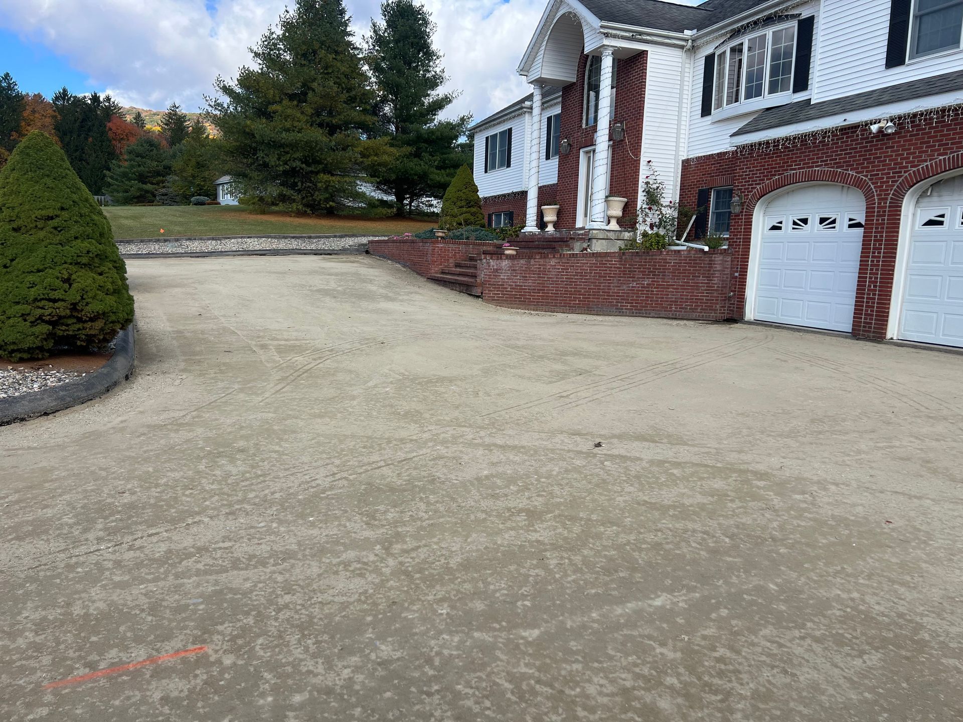 Driveway leading to a two-story house with red brick and white trim. A small evergreen tree is to the left.