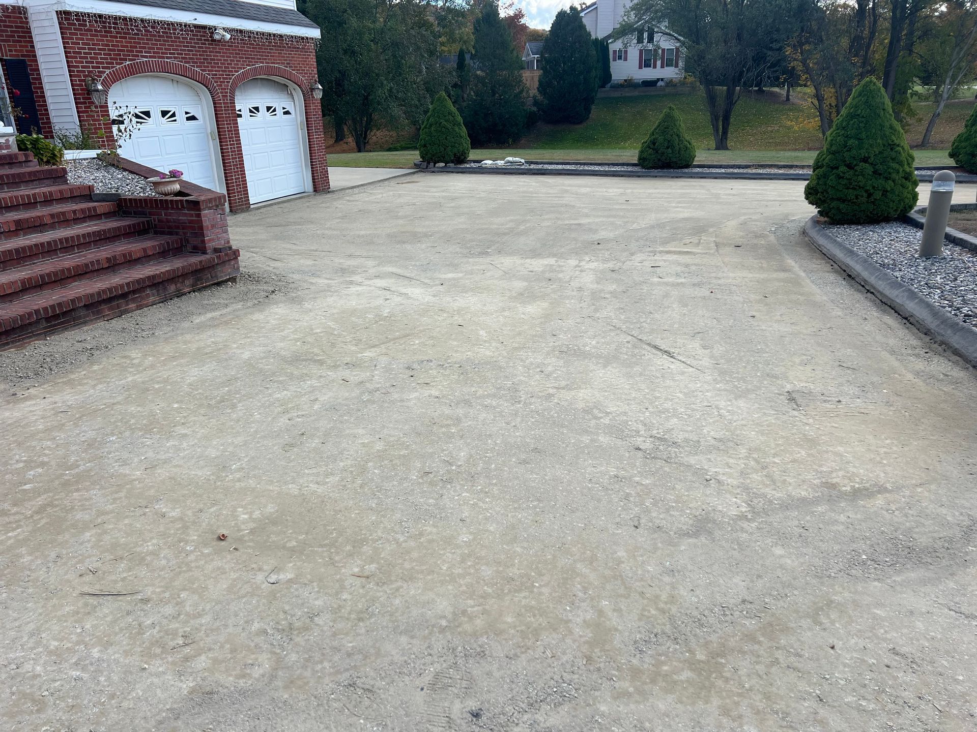 A gravel driveway in front of a brick garage and stairs, with small bushes on either side.