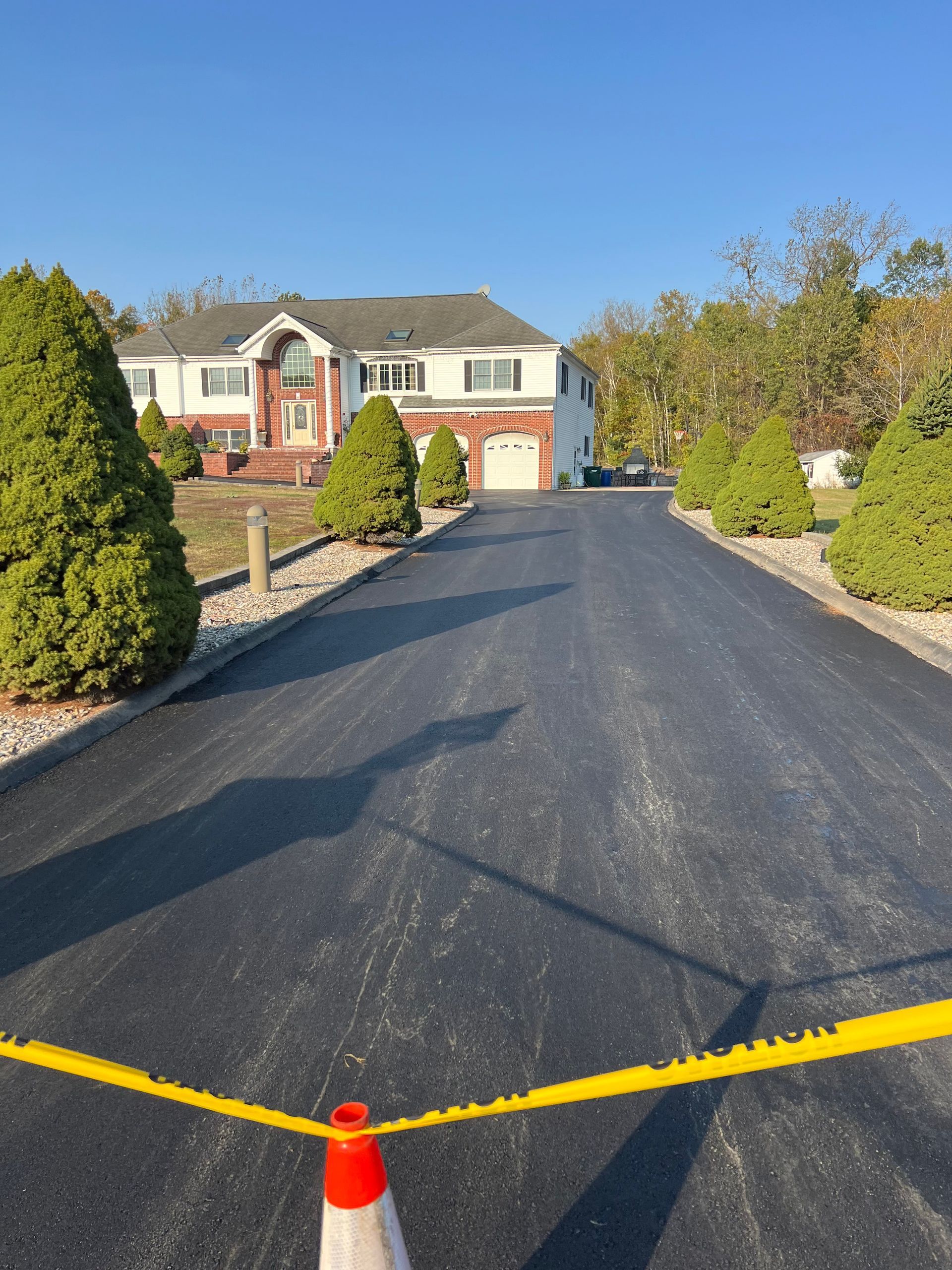 Newly paved asphalt driveway leads to a two-story house with brick accents. Yellow caution tape is in the foreground.