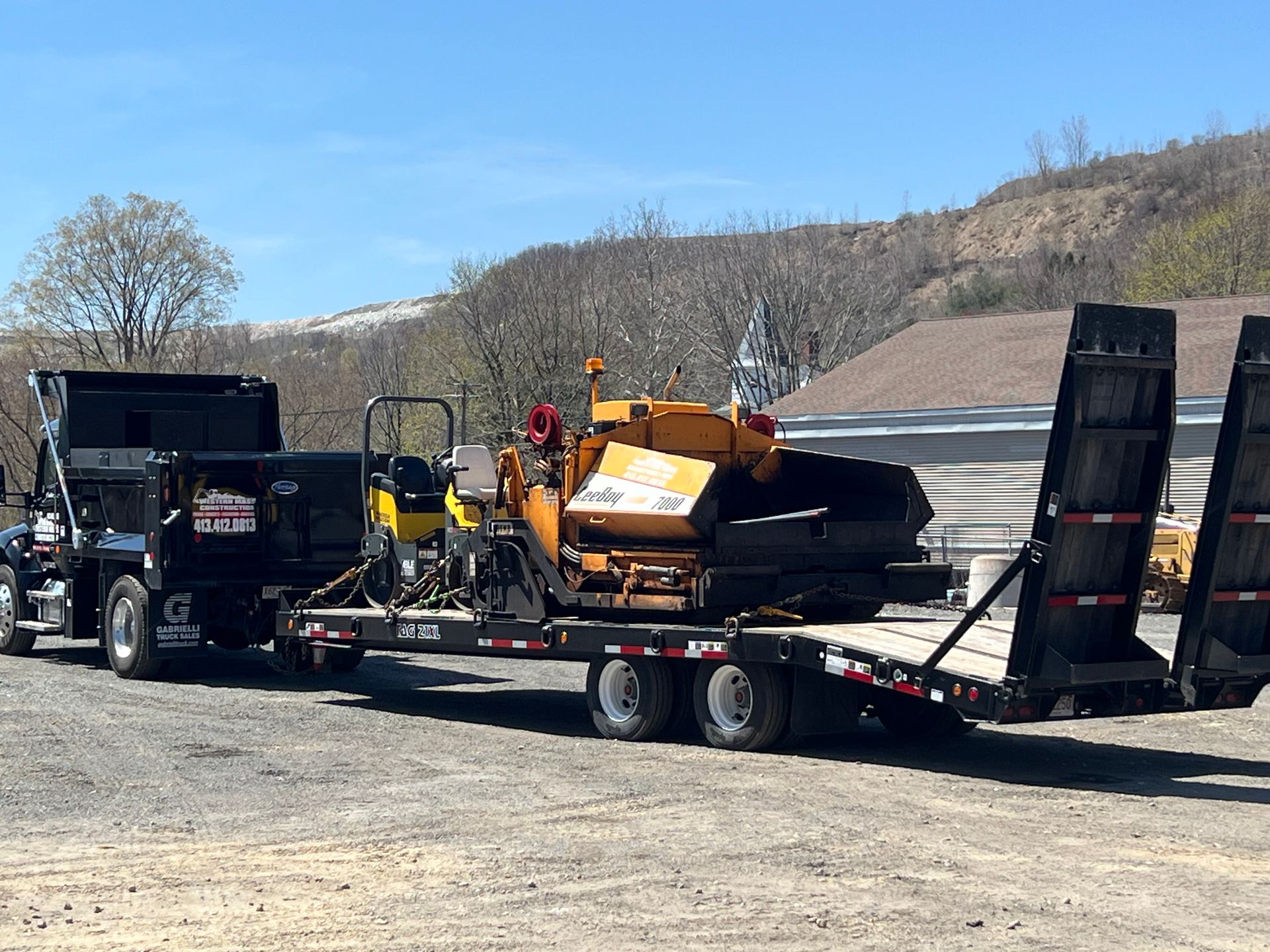 Truck pulling trailer with paving equipment. Sunny day, outdoors.