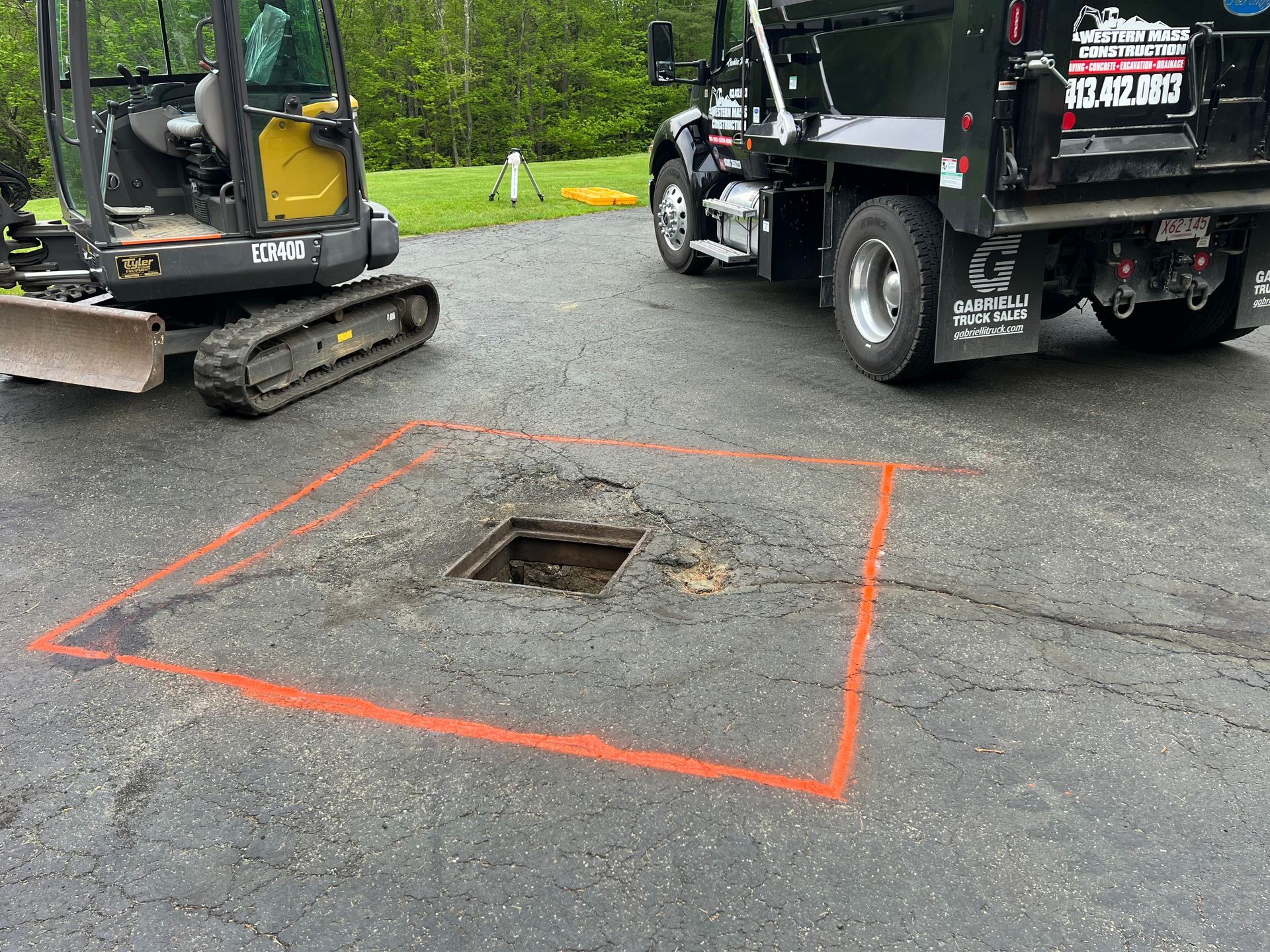 Asphalt with orange square, open utility hole, excavator and dump truck.