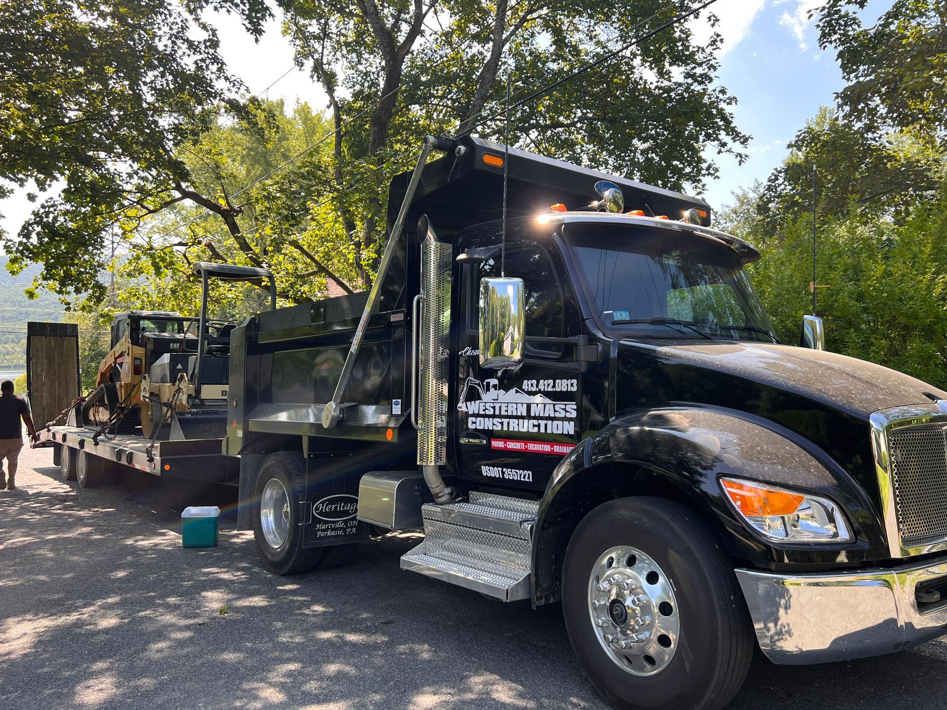 Black dump truck with trailer carrying construction equipment. Outdoors, sunny day.
