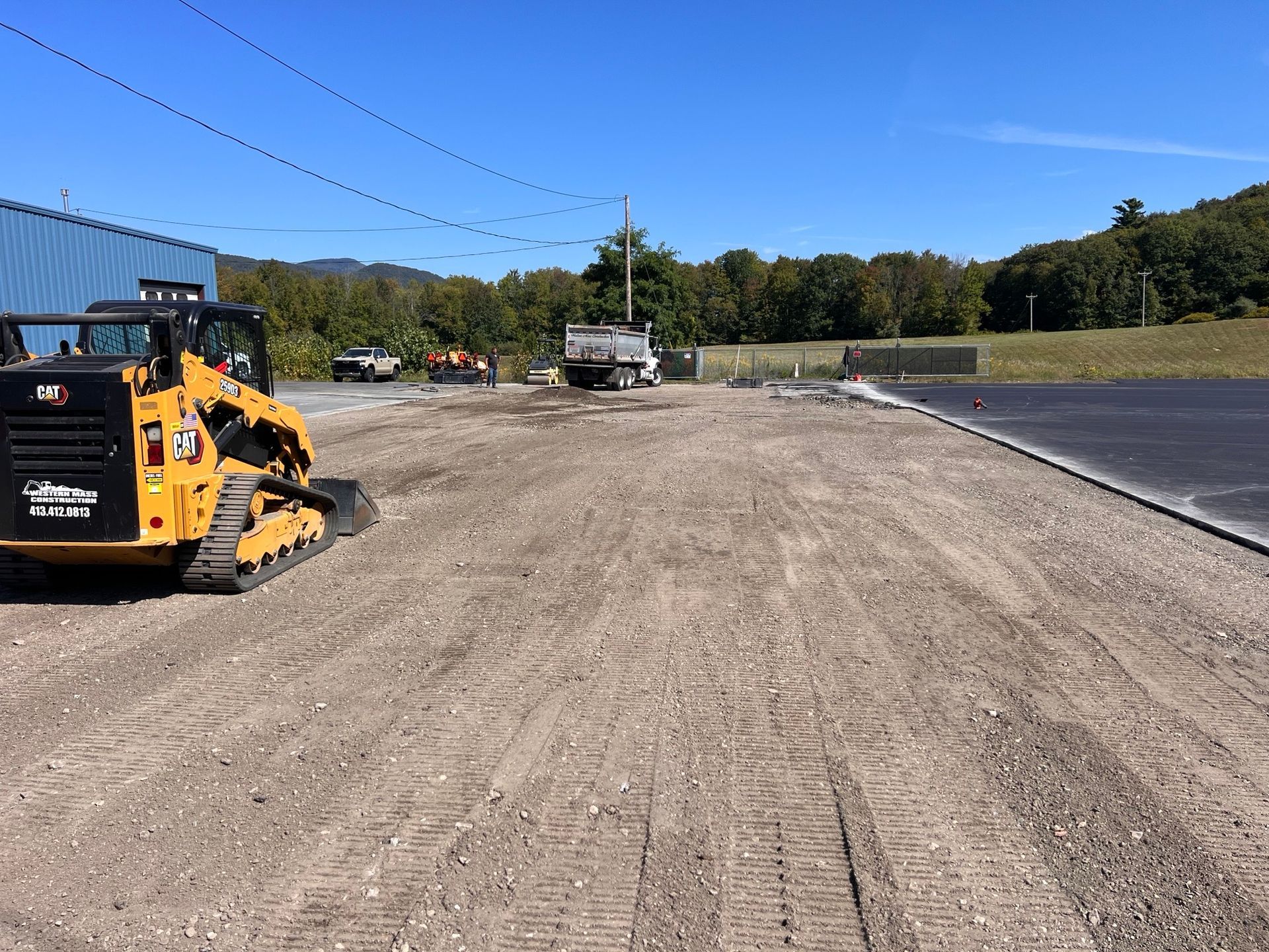Yellow skid steer on gravel, construction site; truck in background. Clear sky, trees.