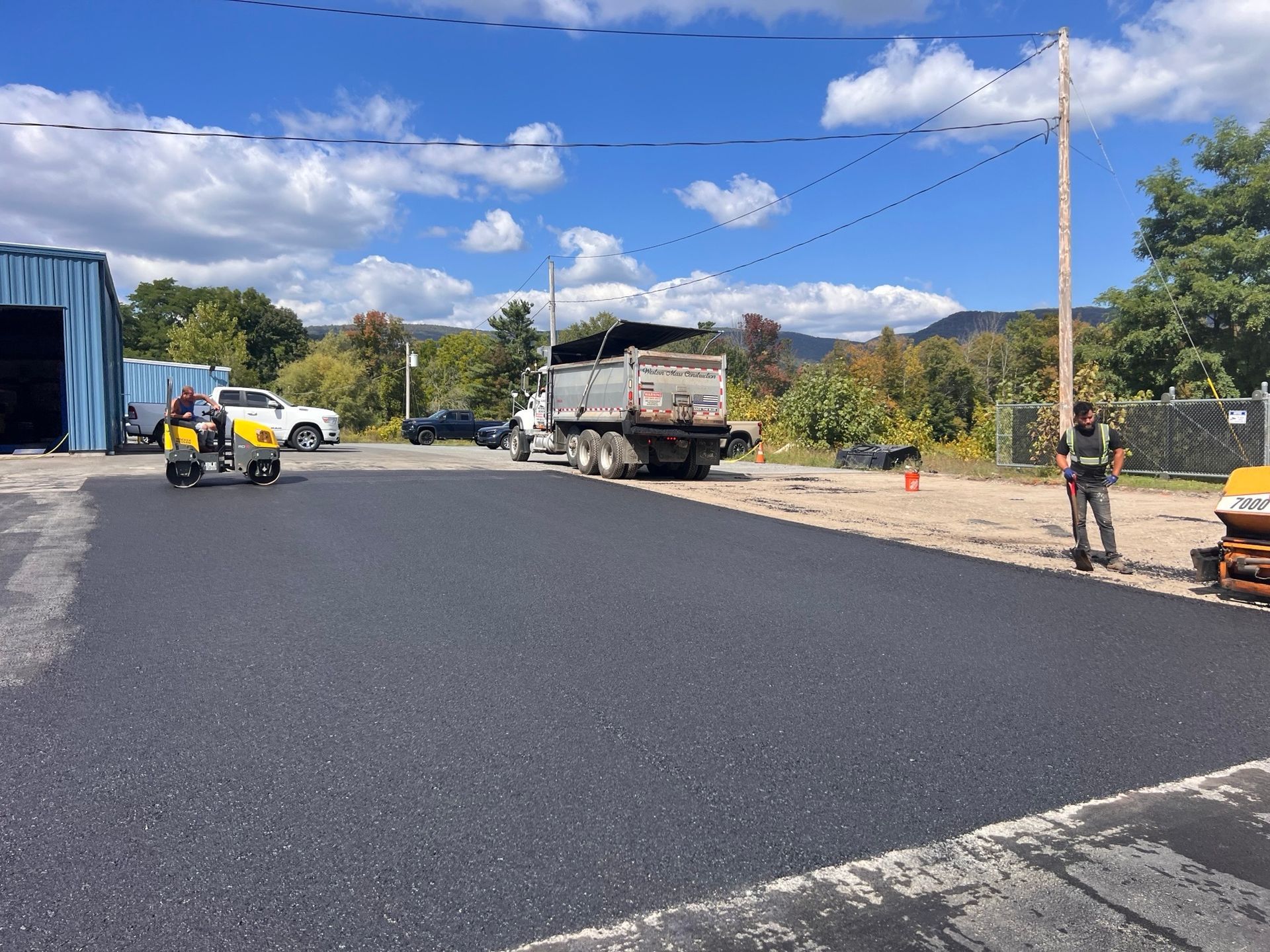 Asphalt paving in progress: truck unloading, workers, compactor, freshly paved surface. Sunny day.