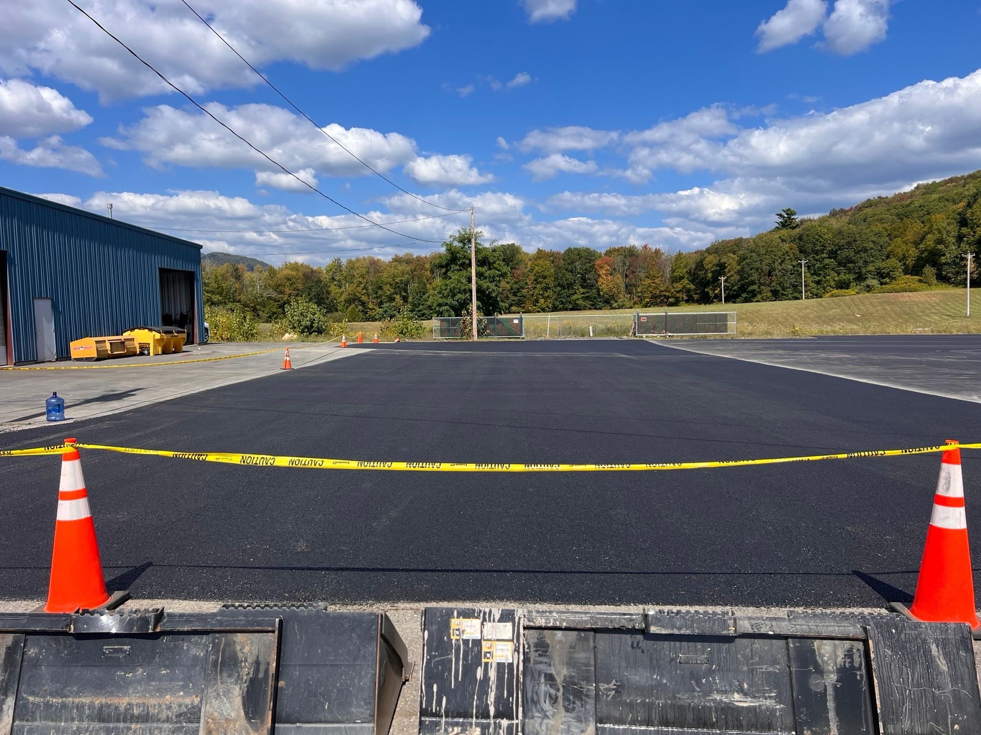 Freshly paved asphalt lot, orange cones and caution tape. Blue building and hillside in background.
