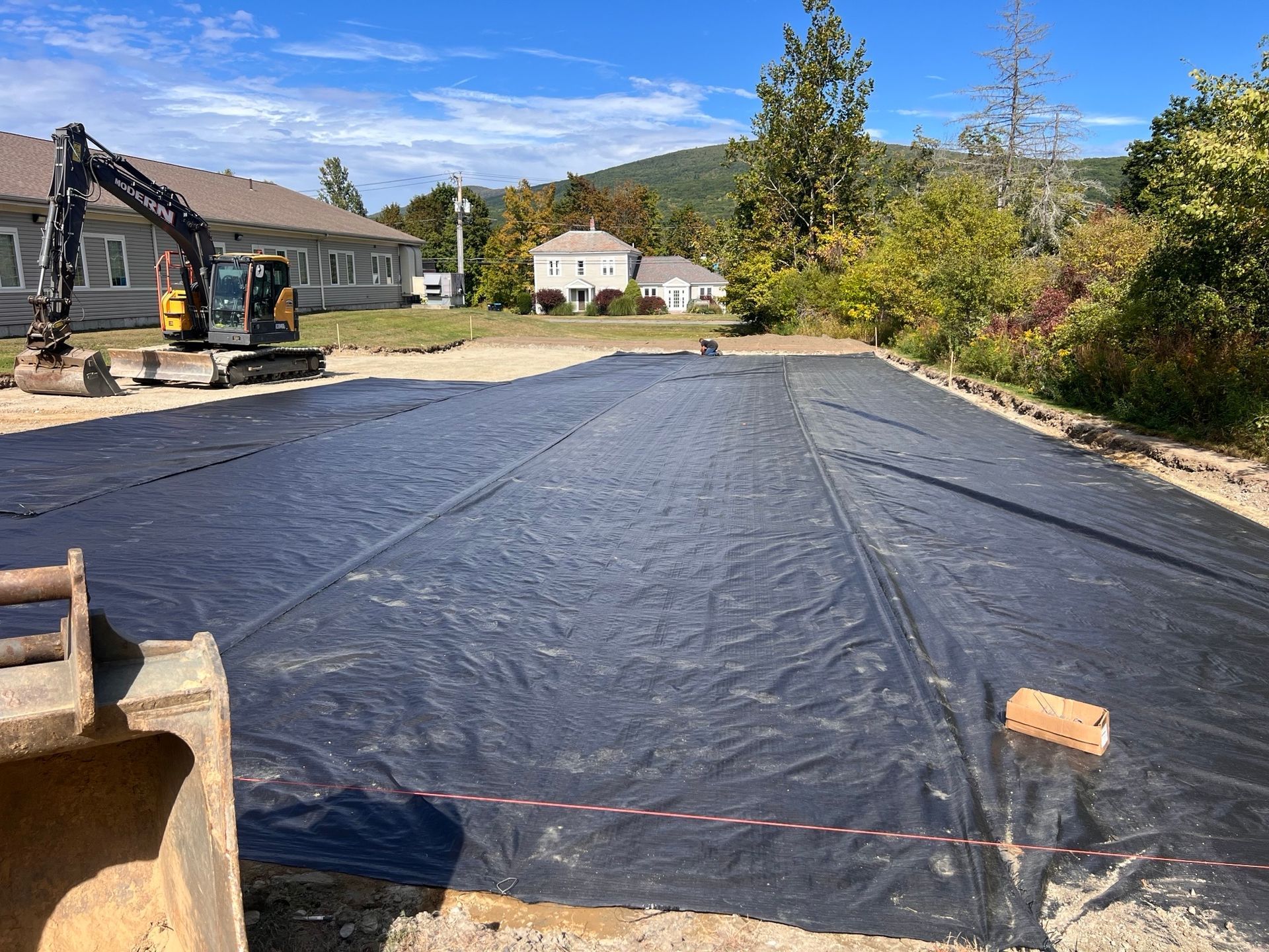 Black landscape fabric covers a cleared area; a small excavator is visible on the left.