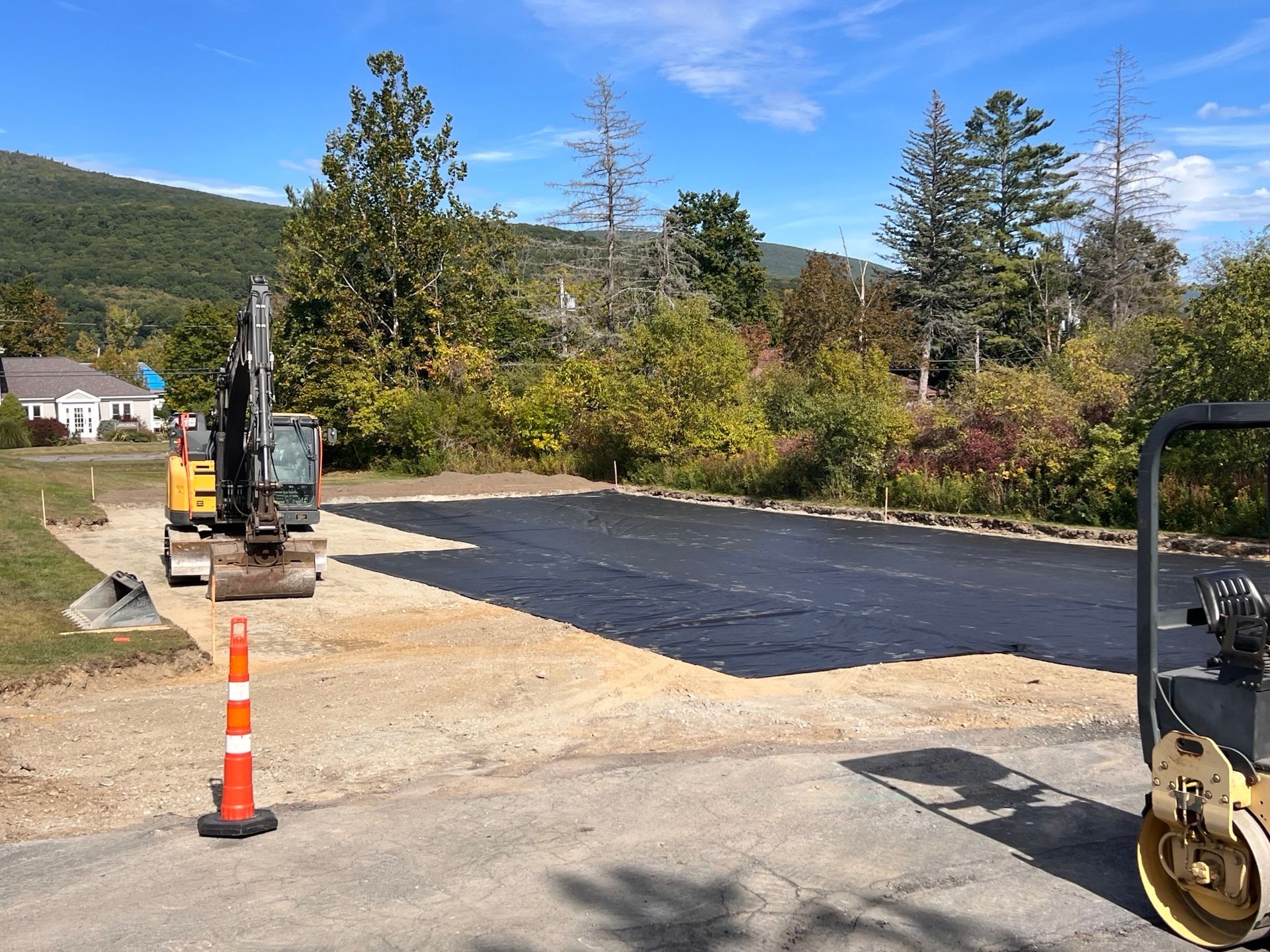 Asphalt being laid in a gravel area; a small excavator and a roller machine present. Green trees and a mountain in the background.