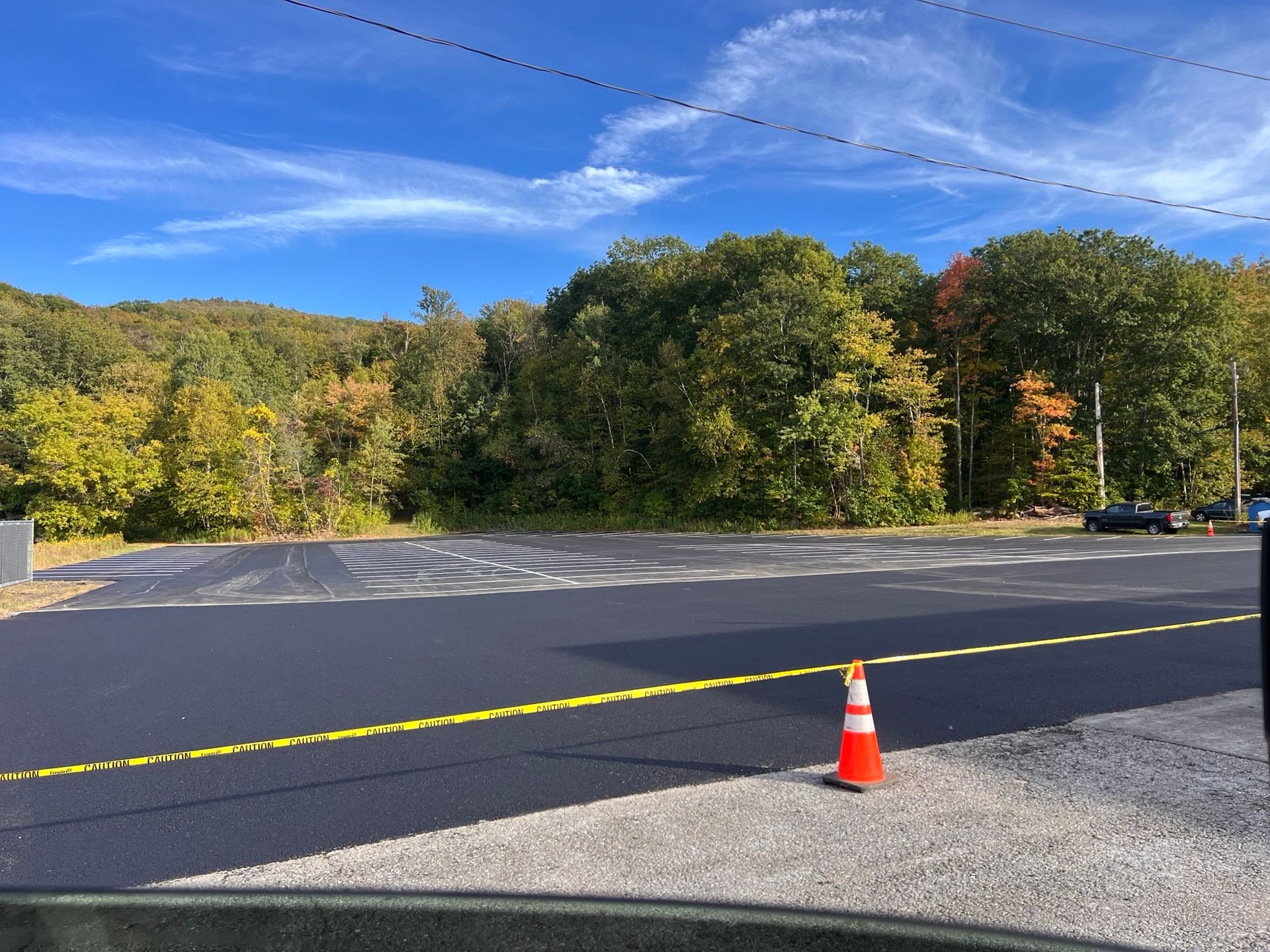 New asphalt parking area with caution tape, traffic cone, and trees on a hillside under a blue sky.