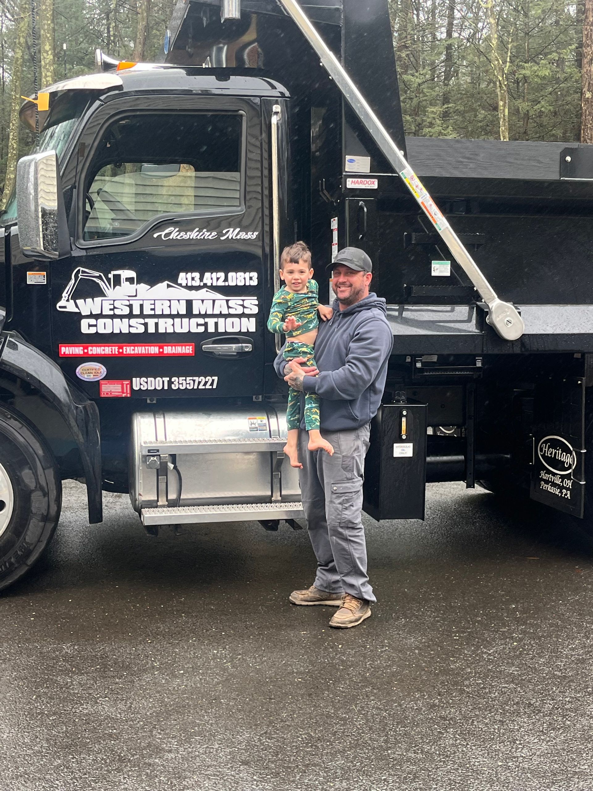 Man holding a child posing next to a black dump truck; they are in front of a construction business.