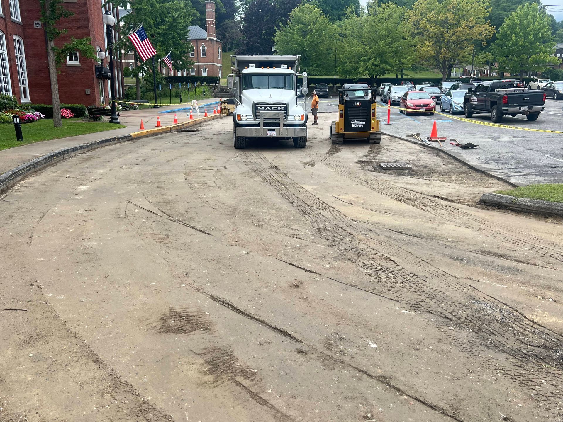 Road construction: Truck and skid-steer loader on a dirt road. Orange cones, brick building, and parked cars visible.