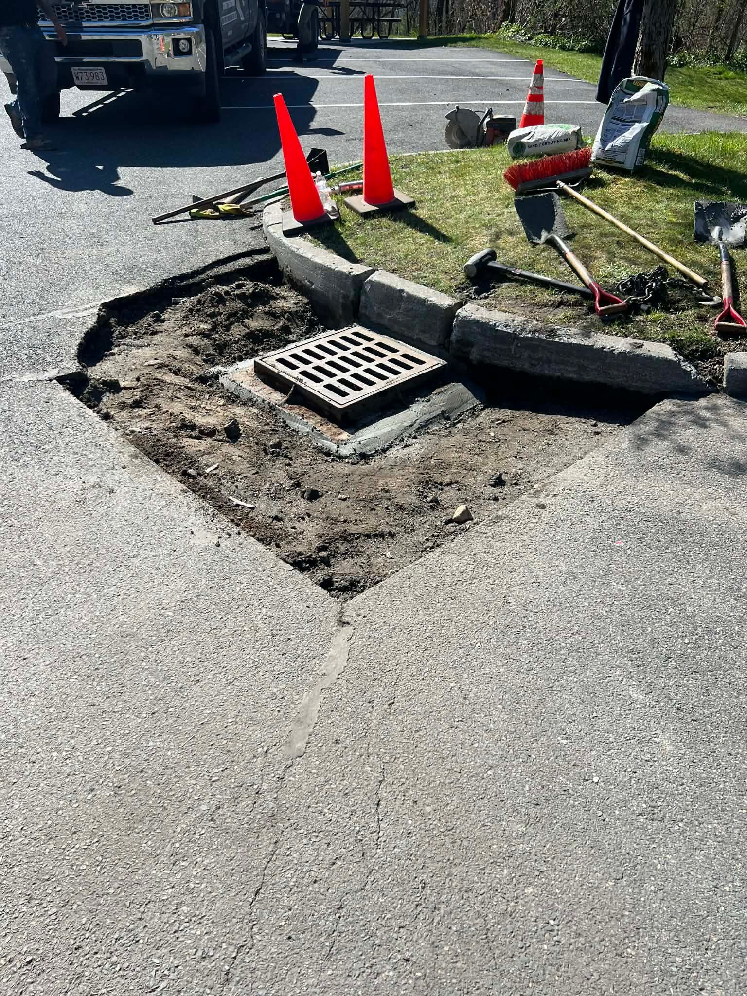 Damaged asphalt around a storm drain being repaired, with traffic cones and tools present.