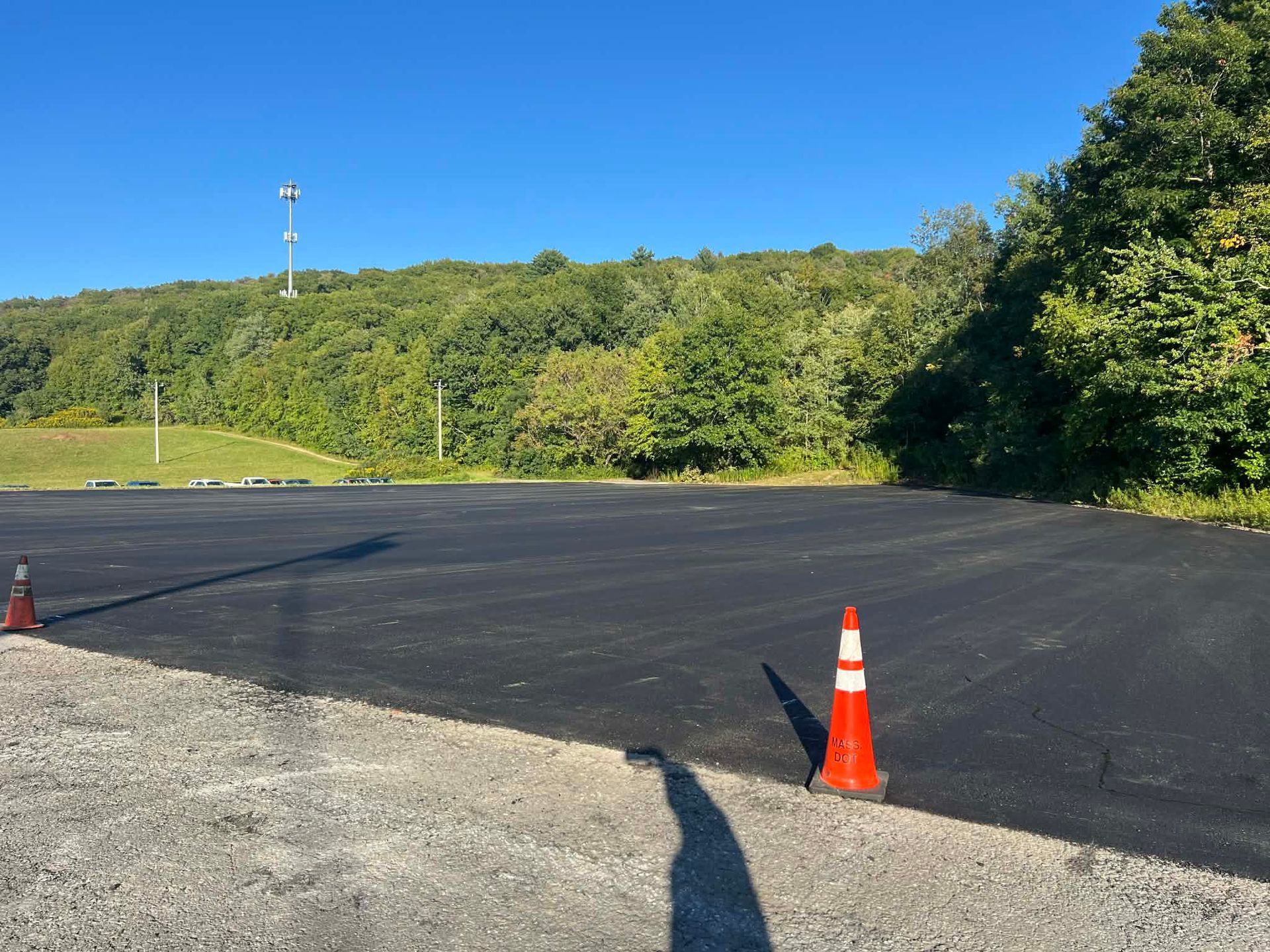 Newly paved asphalt parking lot with orange cones, in front of a treeline and clear blue sky.