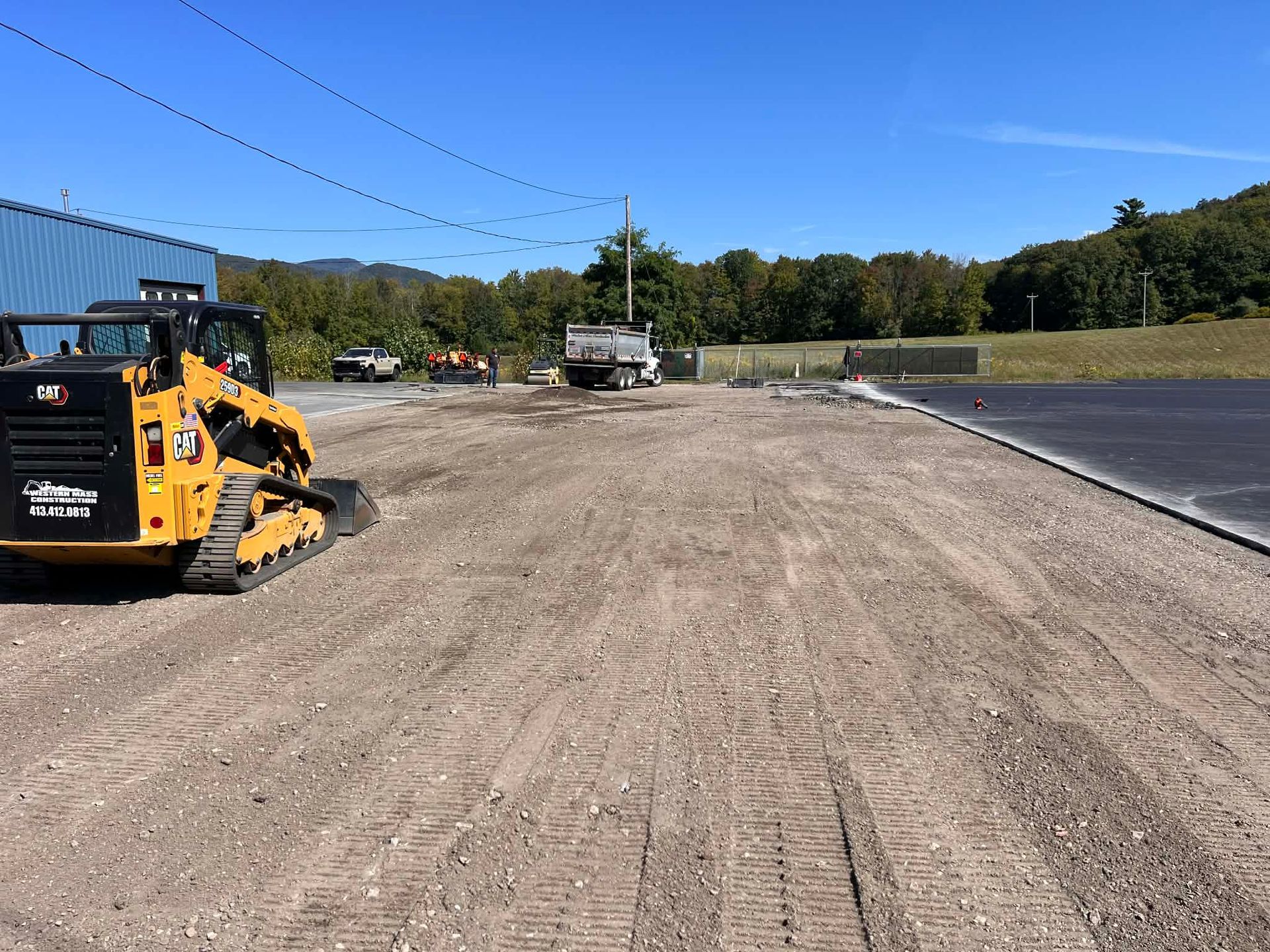 Yellow skid steer on gravel. Construction site with truck, blue building, and trees.