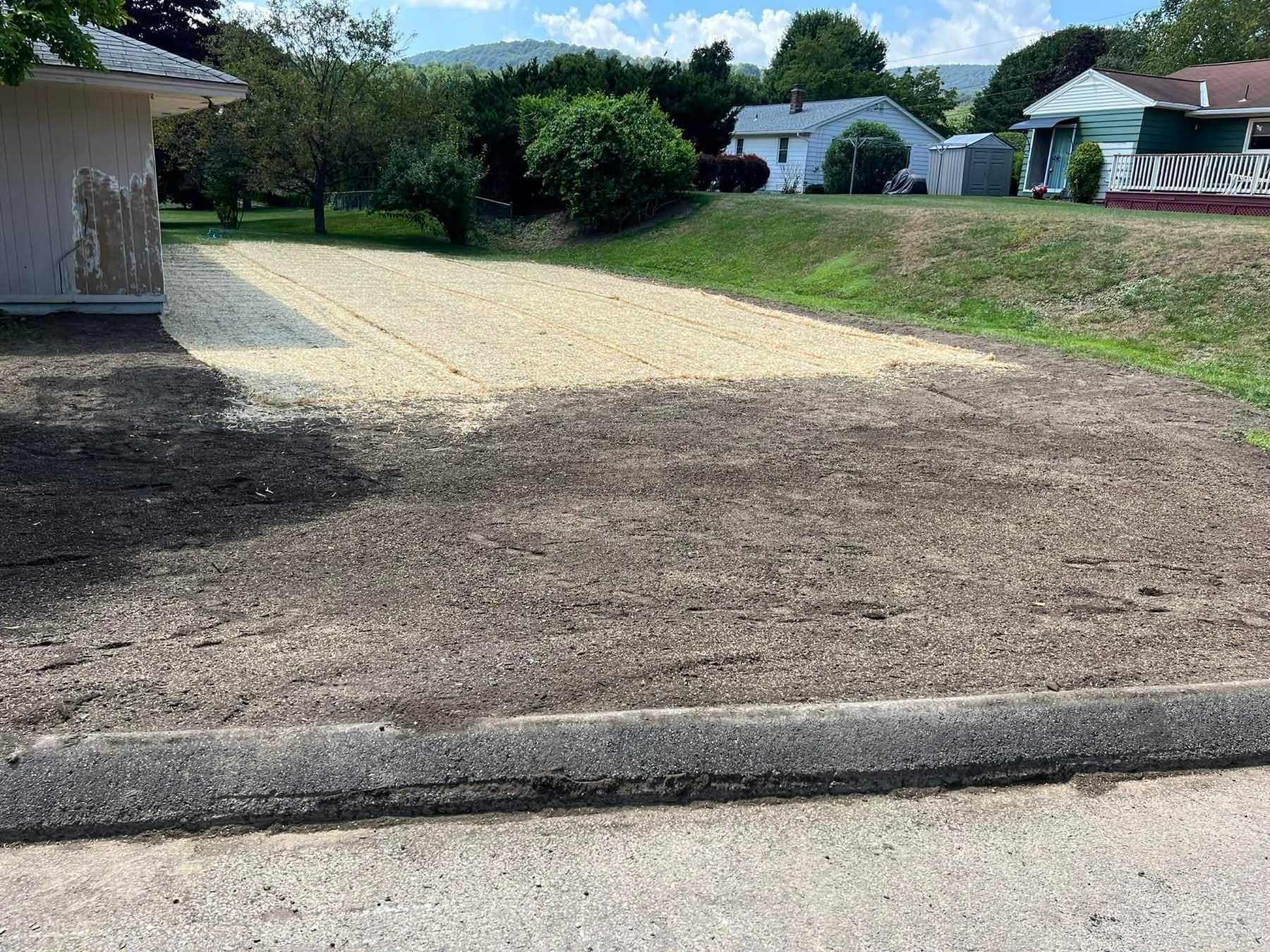 Gravel driveway next to a bare dirt yard, bordered by a concrete curb and houses in the background.