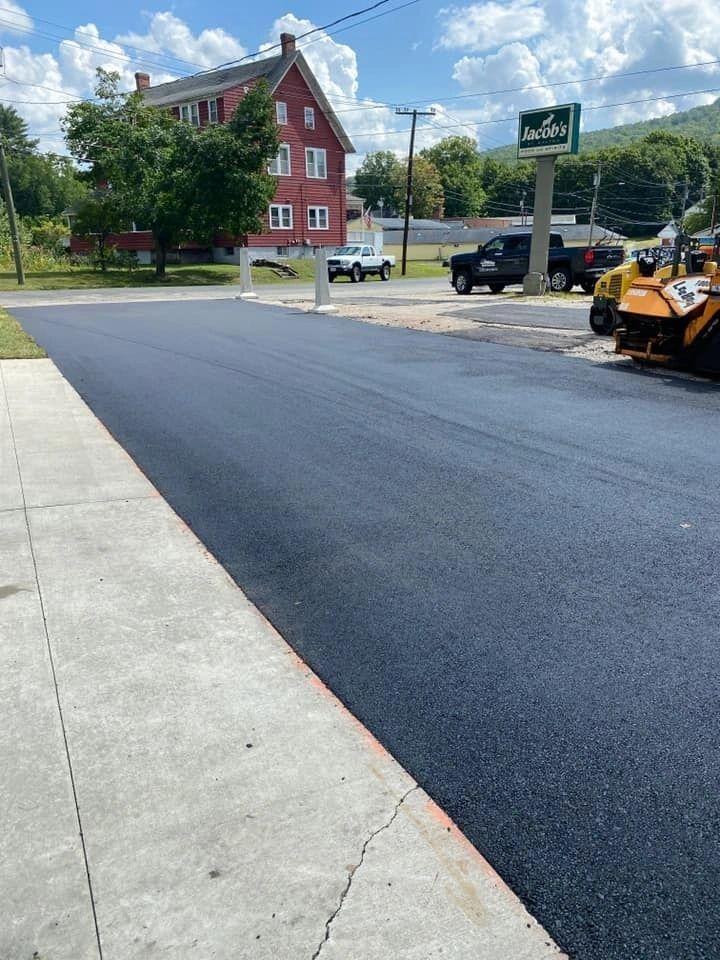 Paved black asphalt road next to a concrete sidewalk, with a red brick building and parked vehicles in the background.