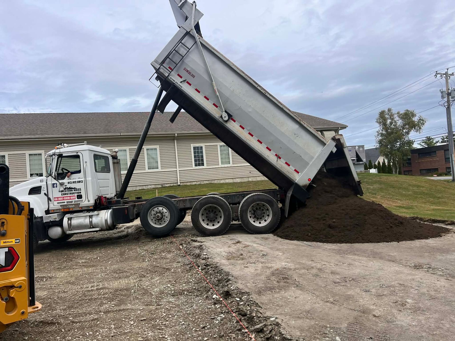 A dump truck is unloading dark soil onto a gravel area in front of a building under a cloudy sky.