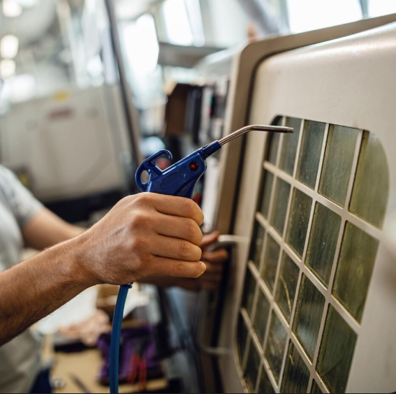 Persona sosteniendo una pistola de aire azul, limpiando una máquina blanca con una rejilla.