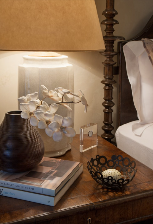 Bedside table with lamp, white flowers in a vase, stacked books, and a decorative bowl beside a bed