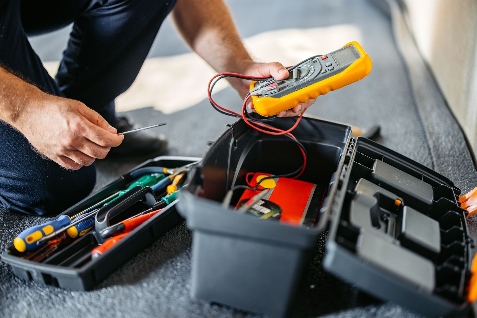 A man is kneeling on the floor holding a multimeter next to a toolbox.