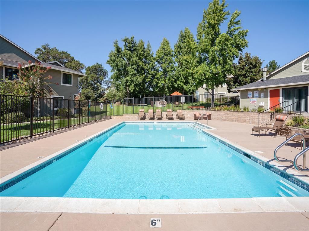 Outdoor community pool with blue water, lounge chairs, fencing, and surrounding trees.