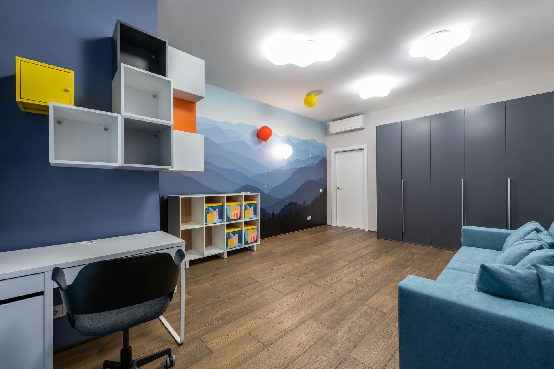 Child's room with desk, sofa, storage, and mountain mural in shades of blue and orange.