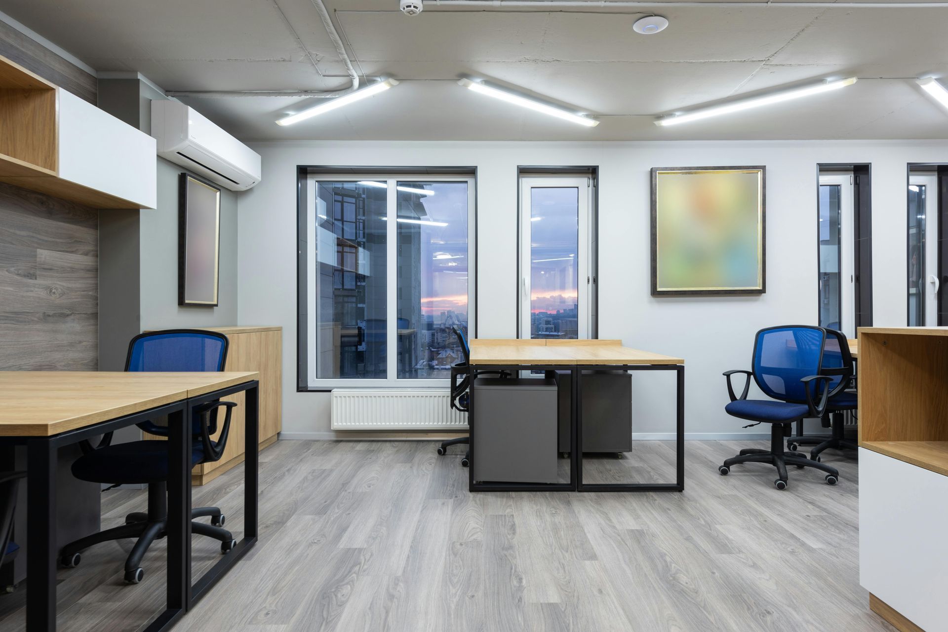 Empty office space with desks, chairs, windows, and artwork. Blue and wooden tones throughout.