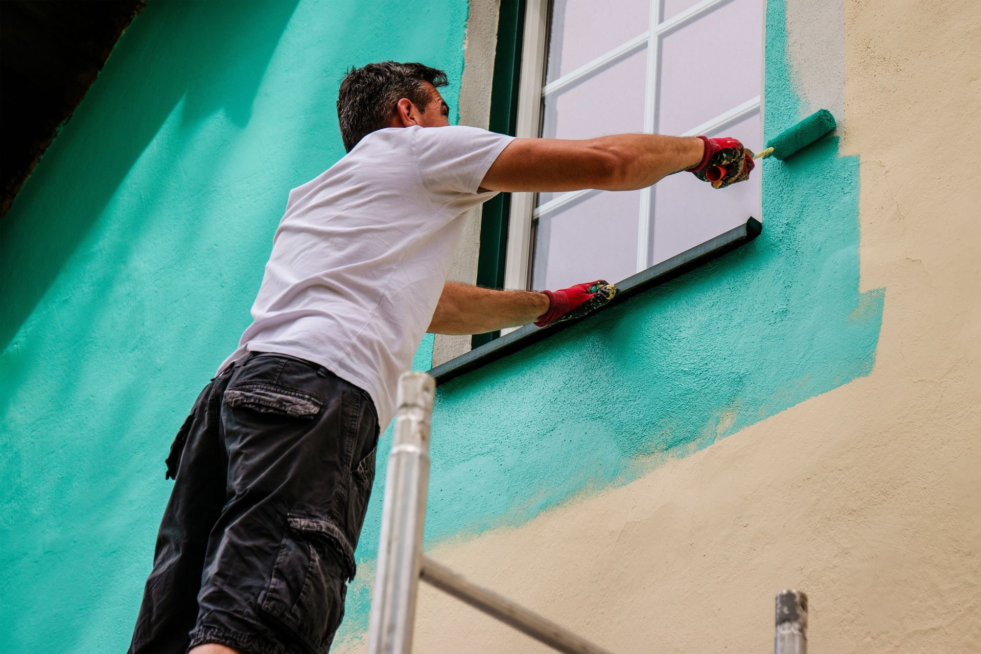 Man paints exterior wall teal, using a roller, next to a window.
