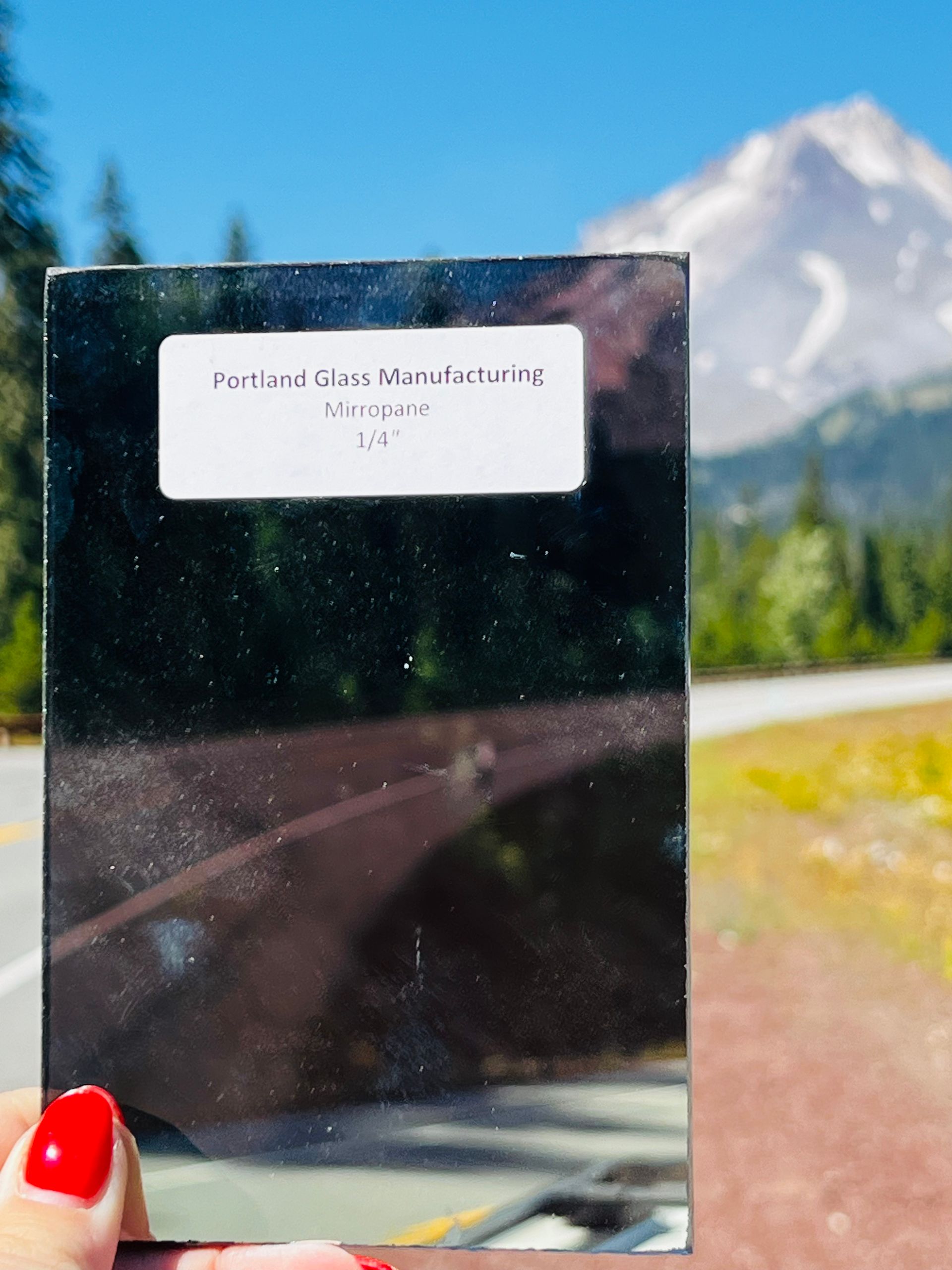 A person with red nails is holding a piece of glass in front of a mountain