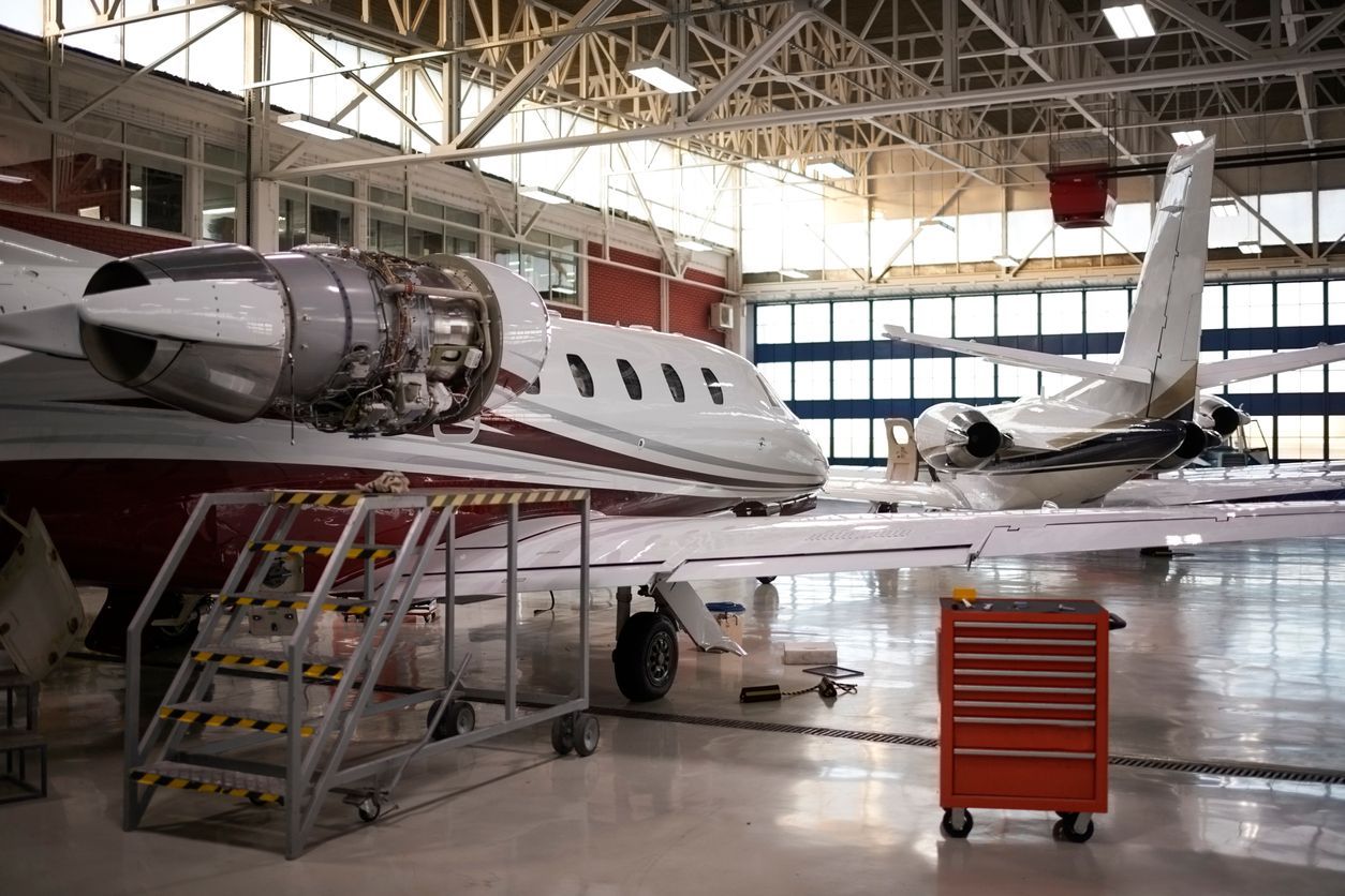Close-up of airplane nose in a hangar; white fuselage, red safety tags, another plane in background.