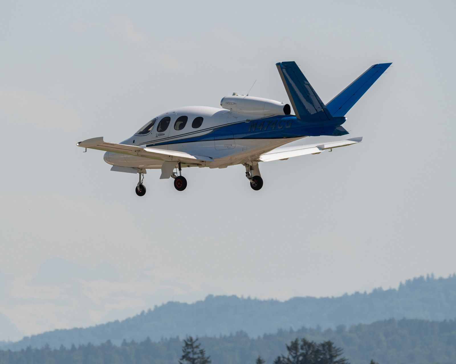 White and blue Cirrus Vision Jet in flight, over a mountain range.