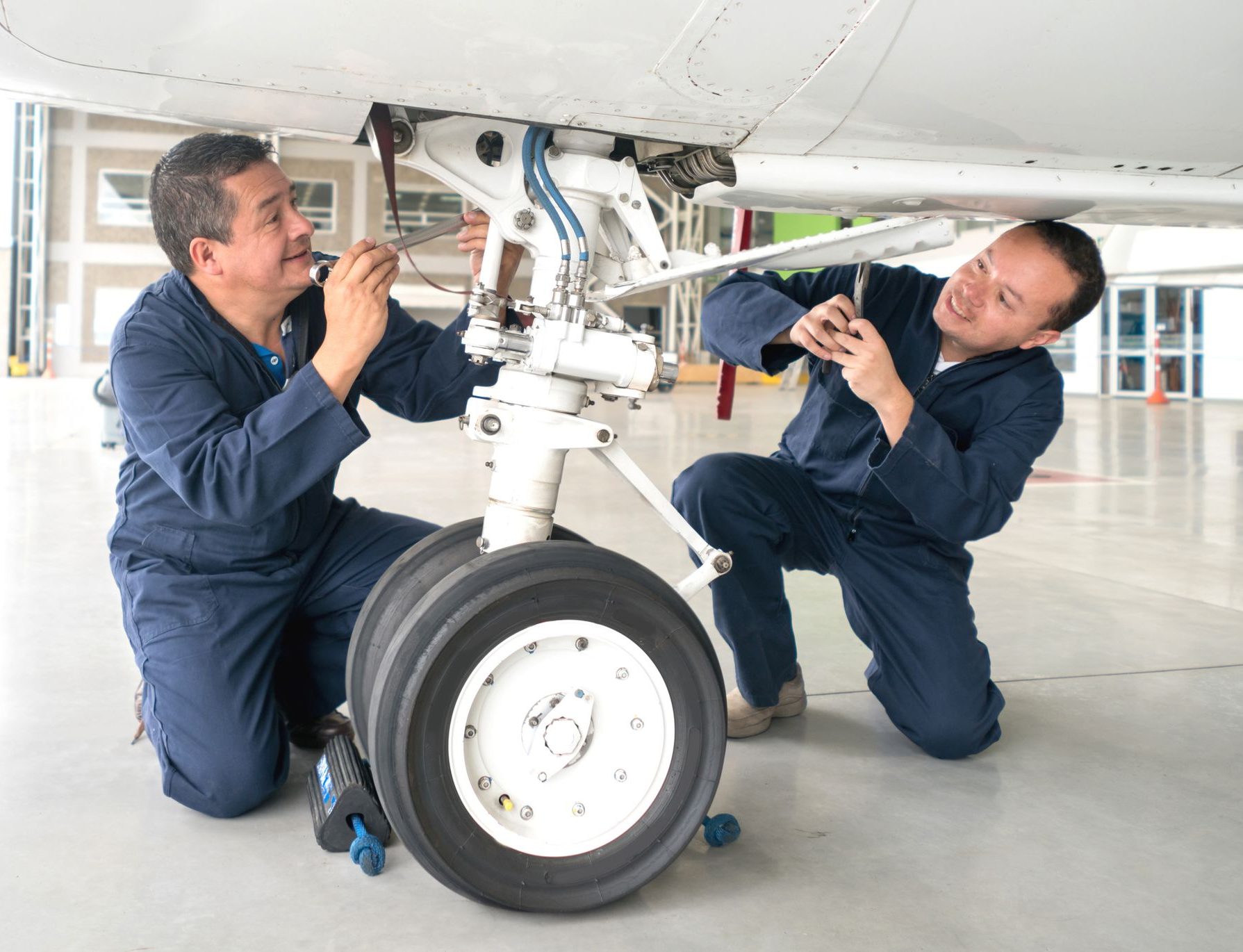 Aircraft mechanic in blue overalls, working on wing underside, using a lift.
