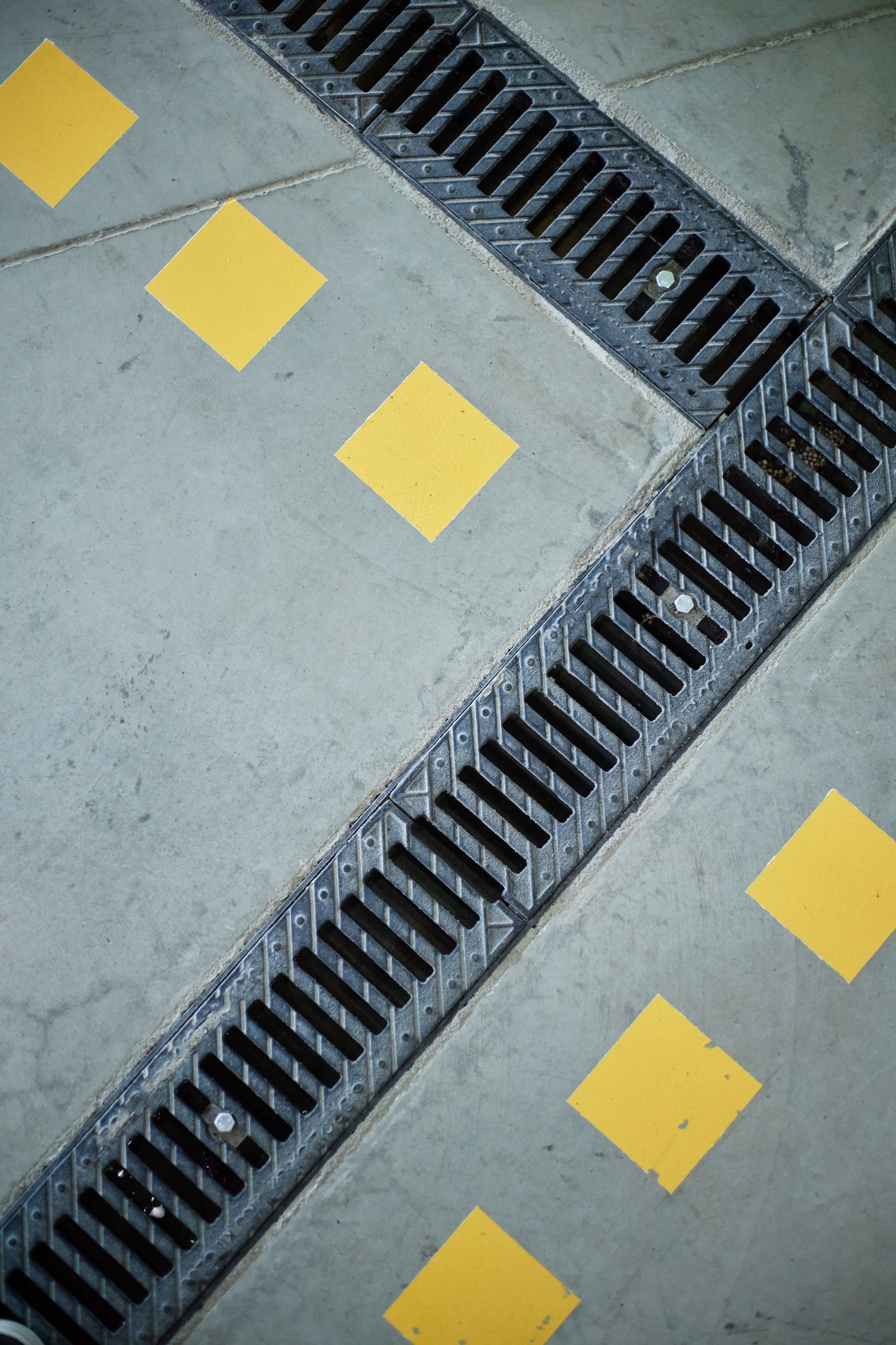 Metal drainage grates intersecting on a concrete surface, with yellow square markers.