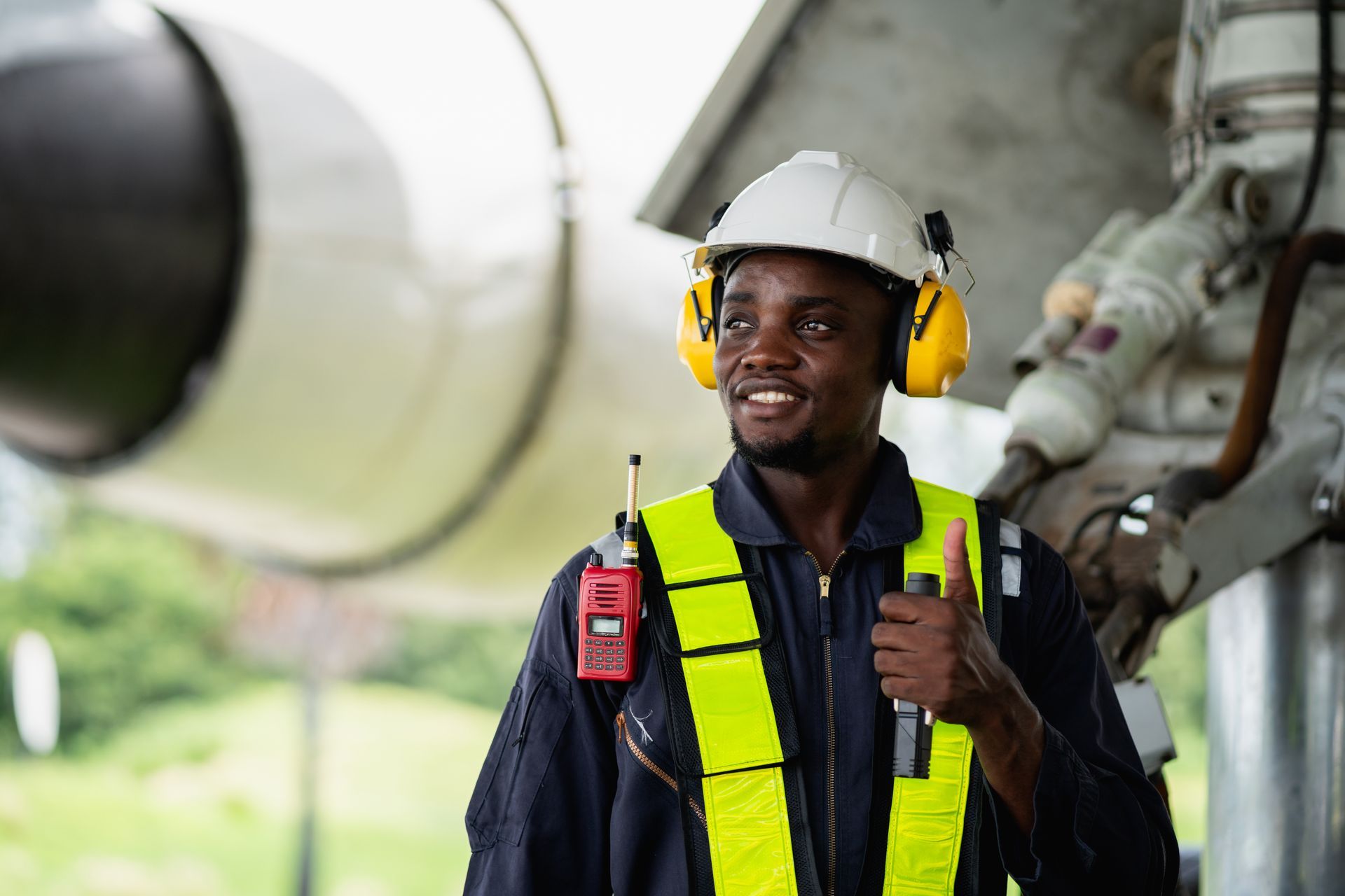 Aviation technician gives a thumbs up. He wears a white hard hat, ear protection, and a reflective vest. Near an airplane.
