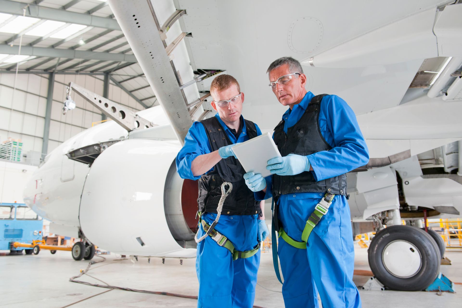 Two mechanics in blue coveralls inspect an airplane, reviewing a tablet in a hangar.