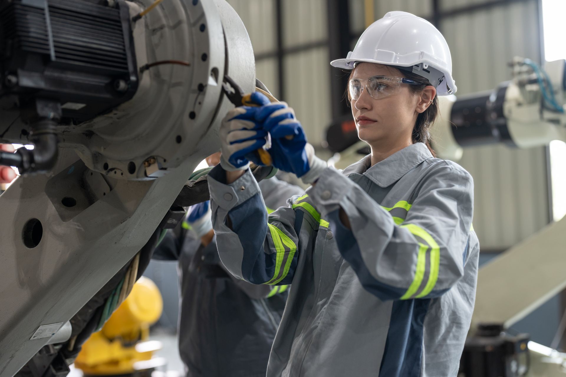 Woman in work attire, wearing safety glasses and a hard hat, works on machinery in a factory.
