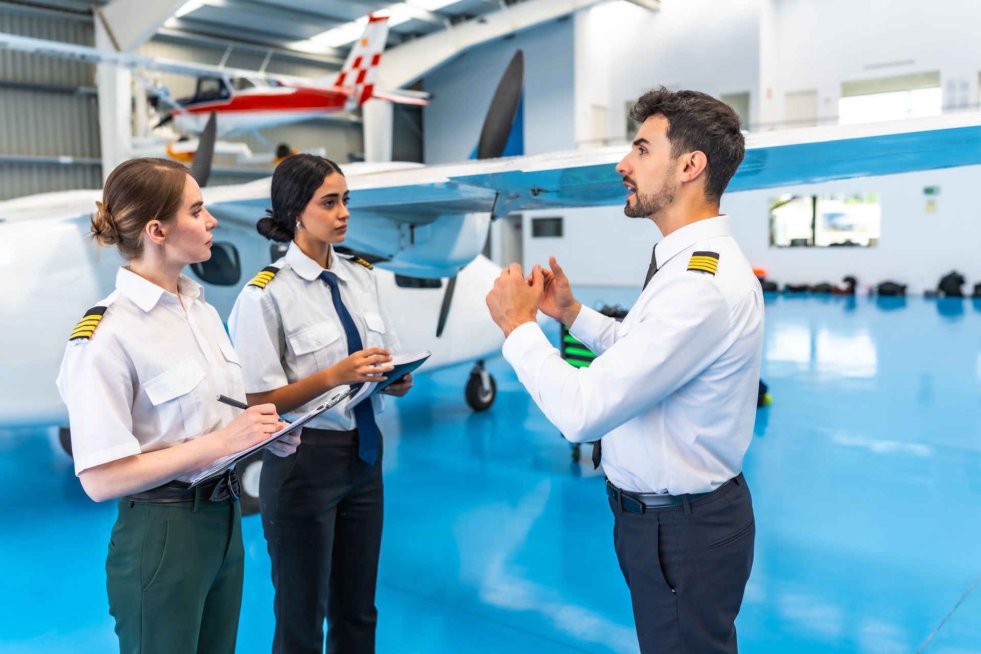 Three pilots in a hangar; one man gestures, two women take notes. White airplane and blue floor.