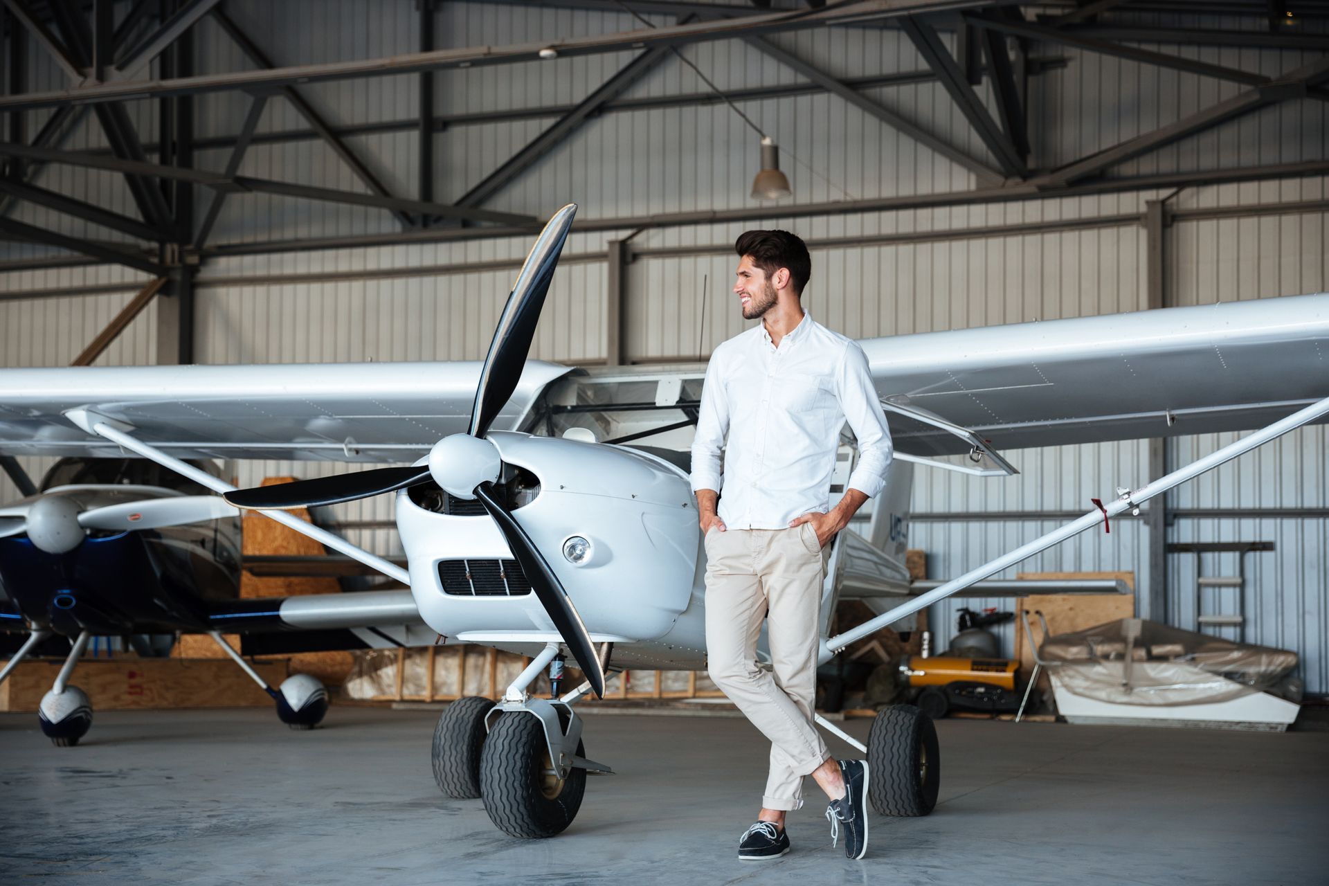 Man stands near small airplane in a hangar, looking away. He wears a white shirt and beige pants.