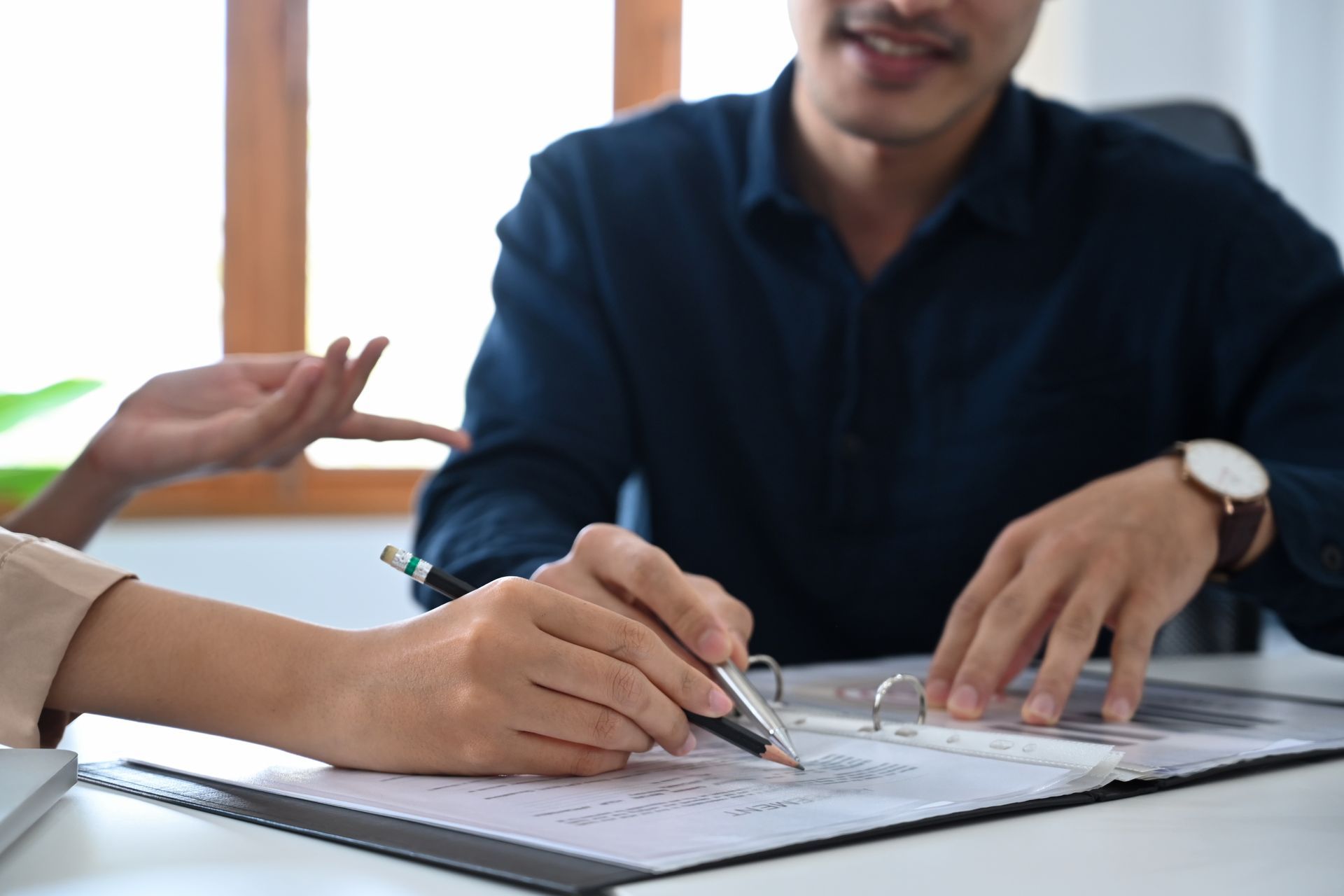 Hands writing on papers in a binder; man smiles while observing; indoor, sunlight.