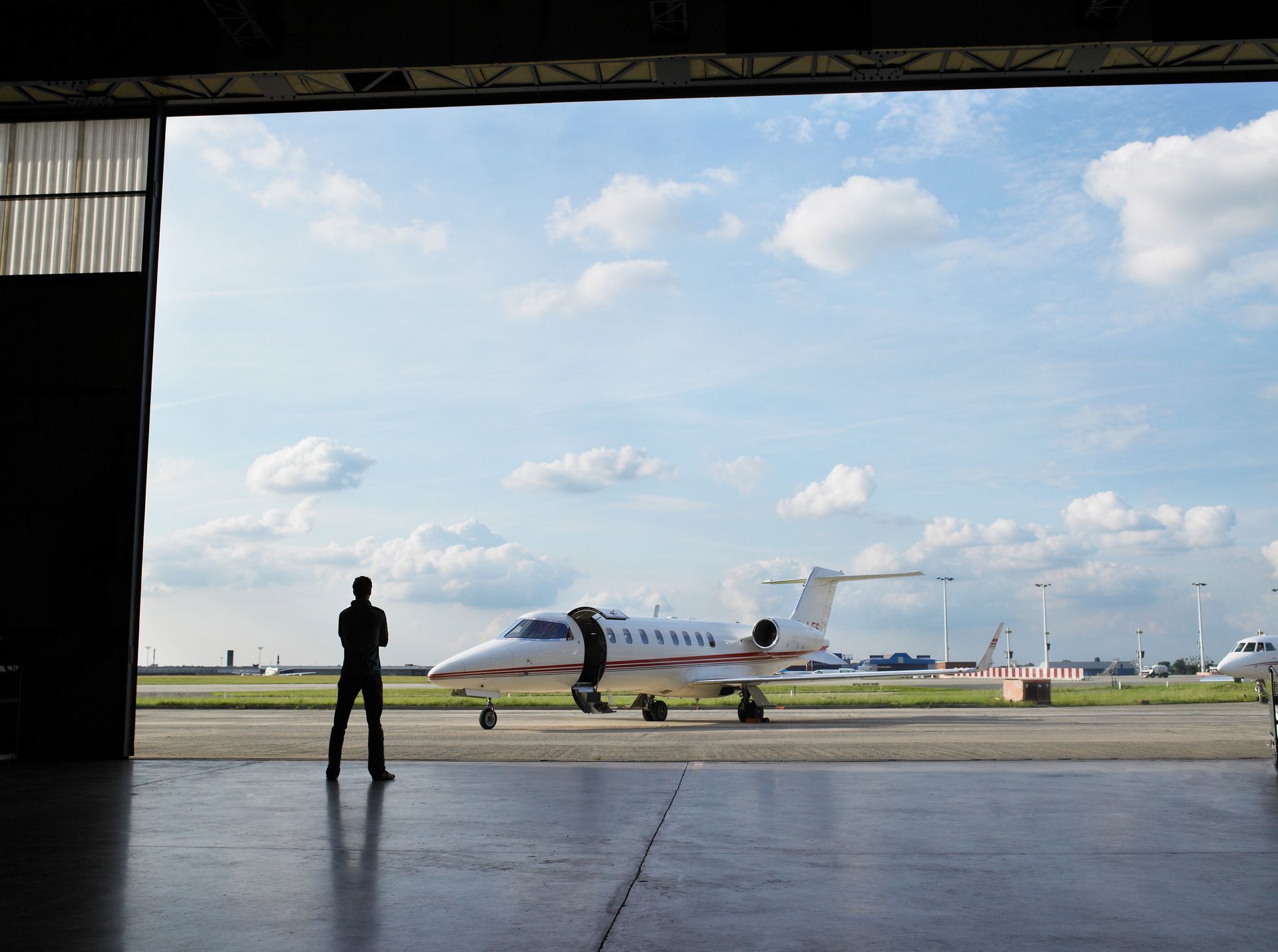 Silhouette of a person standing, looking at a white private jet on an airport runway; visible through an open hangar.