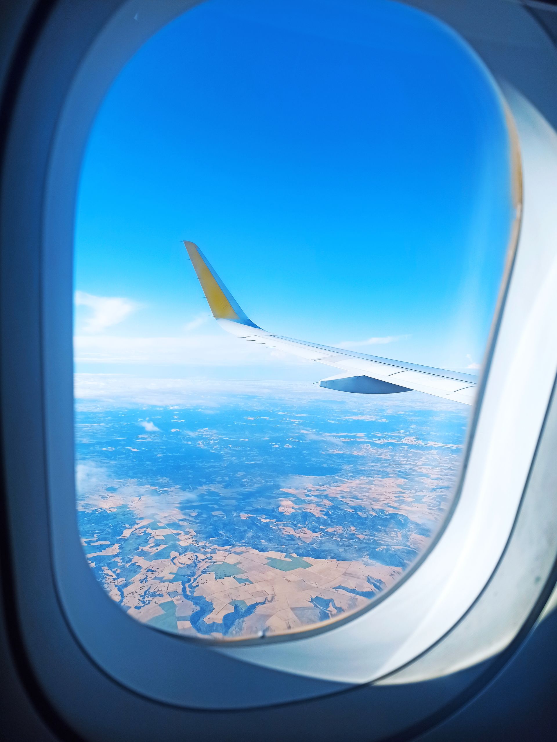 View from an airplane window, showing a blue sky, wing, and landscape with fields.