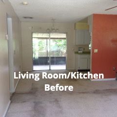 A wide shot of a living room and kitchen interior, featuring beige carpet, an orange wall, and a sliding glass door.