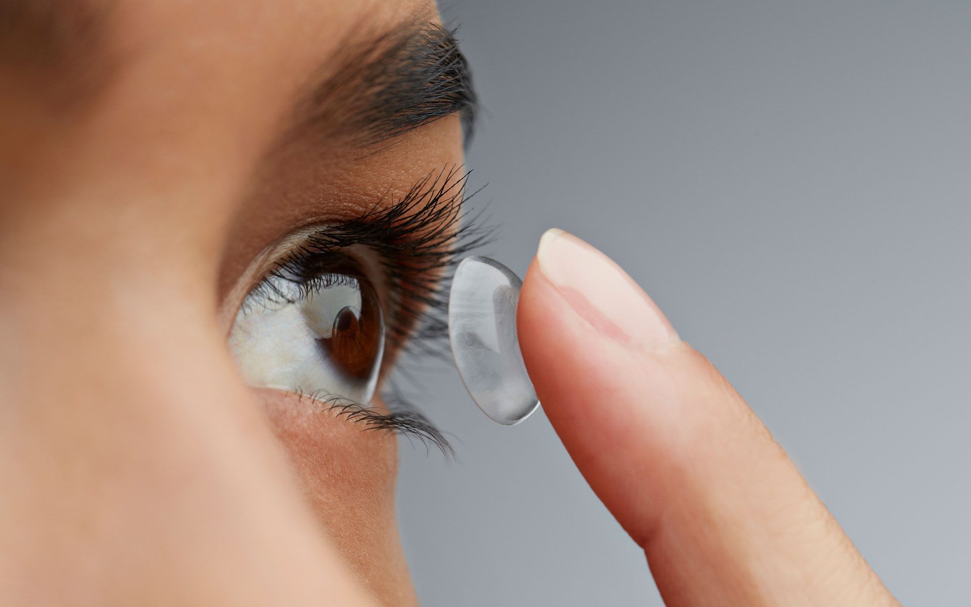 Woman inserting a contact lens into her eye. Brown eye, white skin, gray background.