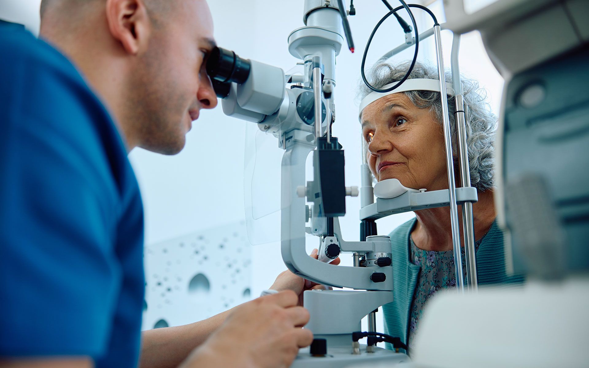 Optometrist examining an older woman's eye with a slit lamp. White medical equipment, blue scrub top.