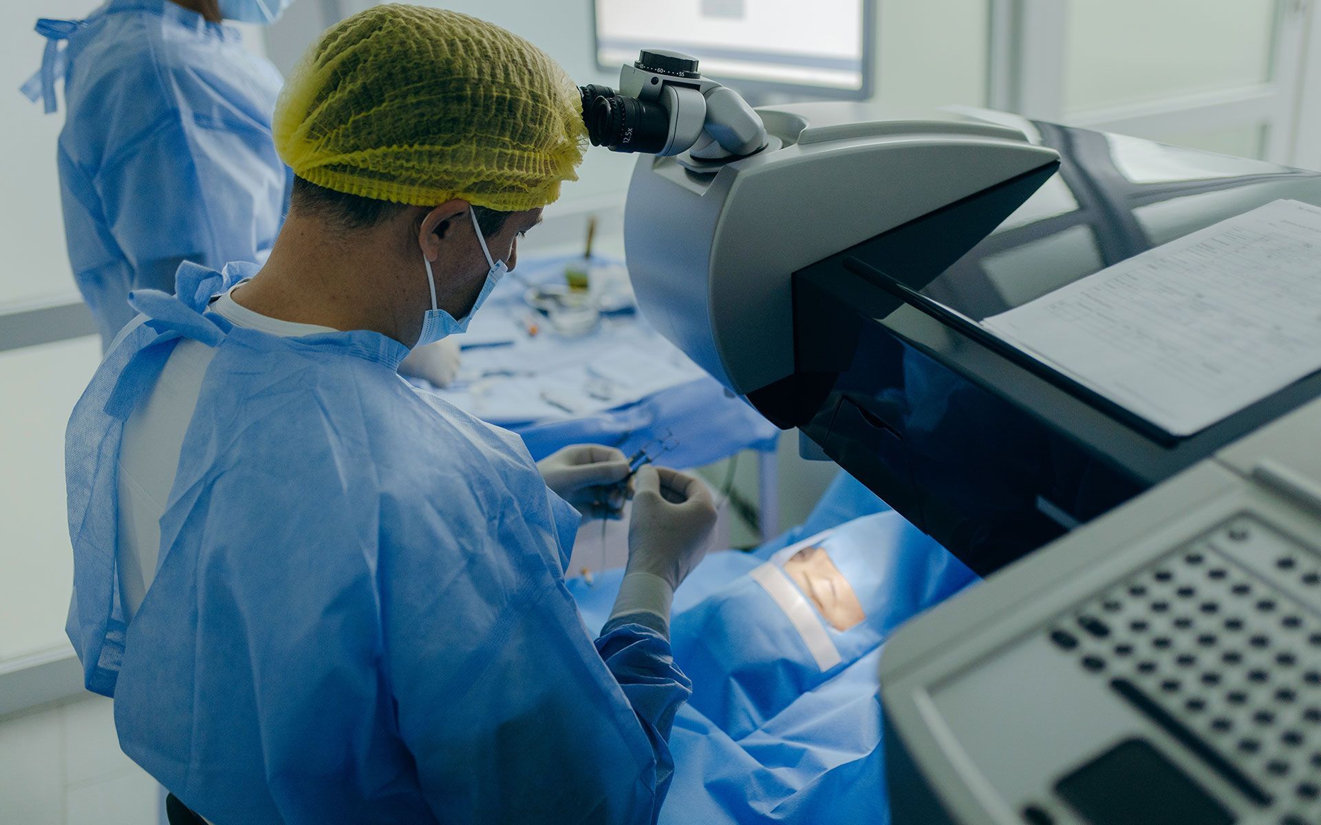 Surgeon in blue scrubs and cap performing eye surgery using specialized equipment.