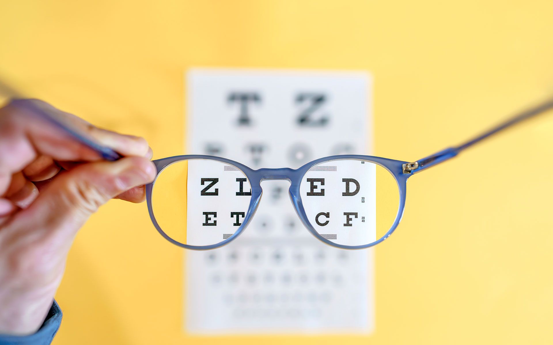 Hand holding eyeglasses over a vision chart on a yellow background.