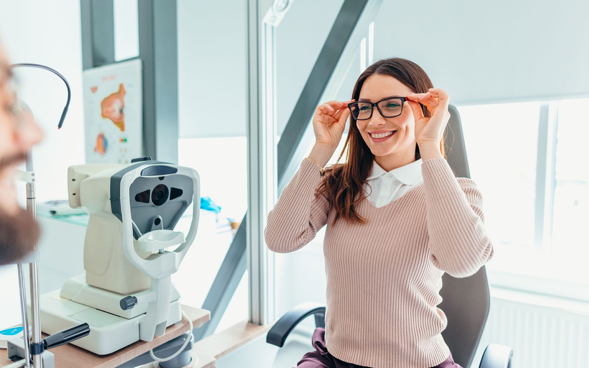 Woman trying on glasses at the optometrist, smiling. Optical equipment in the background.