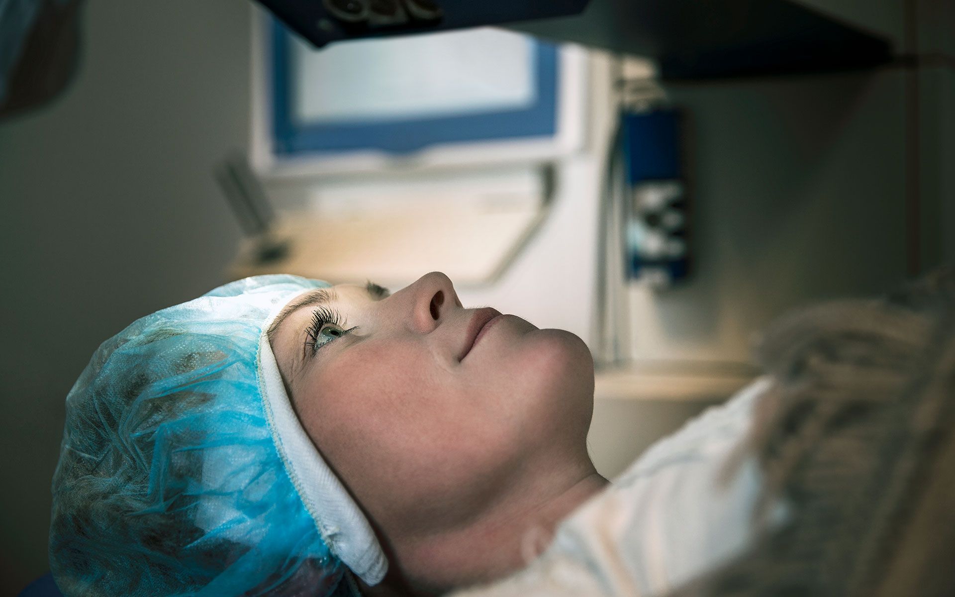 Woman undergoing laser eye surgery, lying down with blue cap, focused expression in medical setting.