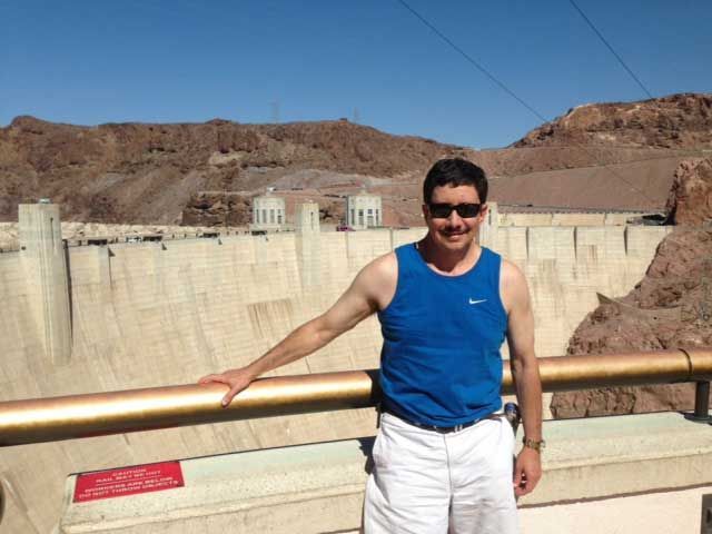 Man in blue tank top and white shorts poses by the Hoover Dam, holding a rail.