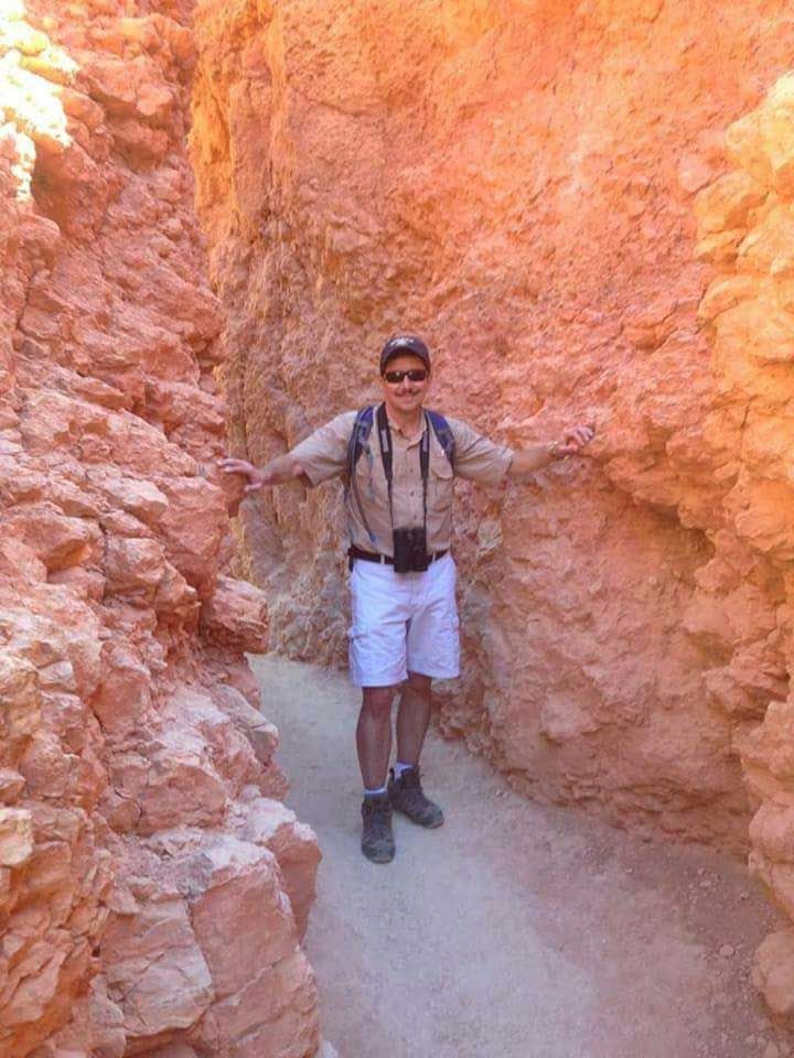 Man with arms out in a narrow red rock canyon. He wears light clothes and sunglasses.