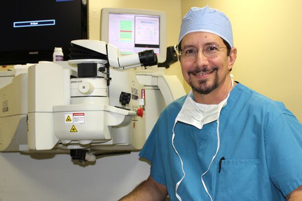 Surgeon smiling in front of eye surgery equipment, wearing scrubs and a cap.